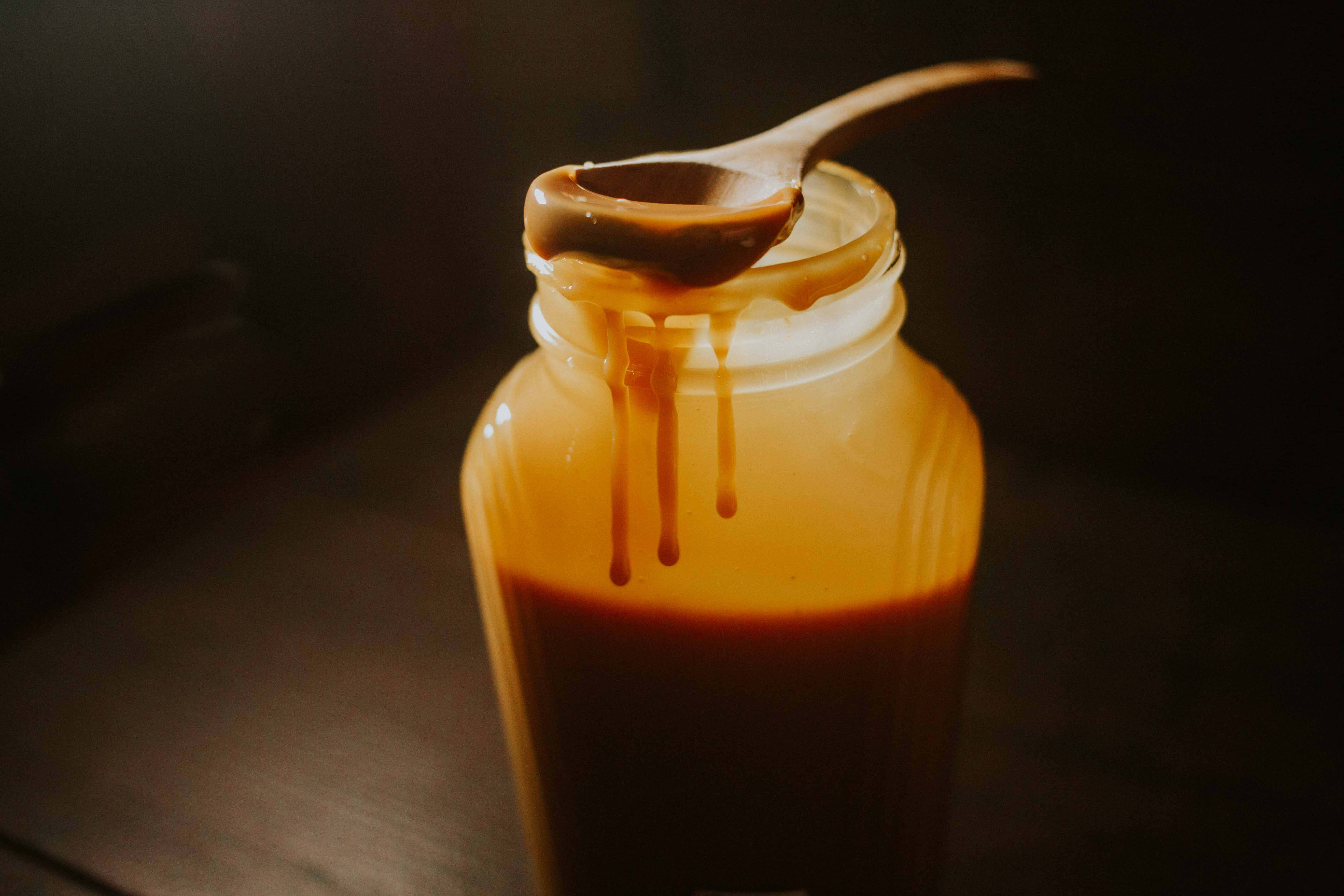 Caramel 桌布 壁紙 — 一罐濃郁的焦糖醬，配以木勺在深色背景下滴下金色糖漿。