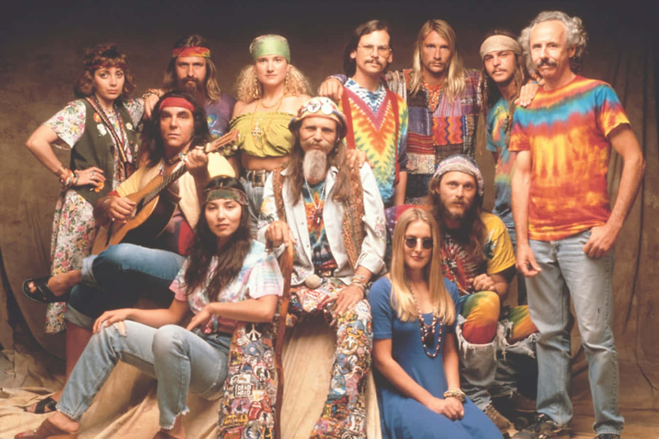 嬉皮士 桌布 壁紙 — 一群11人身穿1970年代嬉皮士服裝，在素色背景前一起擺姿勢，穿著色彩繽紛的紮染襯衫和頭帶。