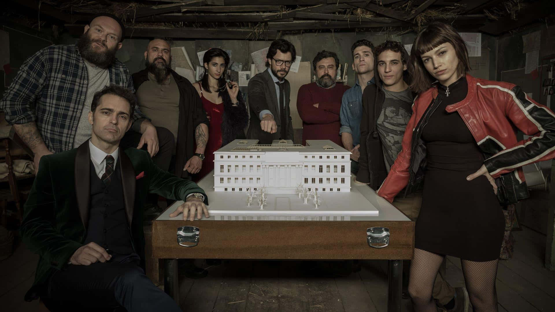 Money Heist 桌布 壁紙 — 一群不同背景的人圍繞著木桌上的一個白色建築模型，身處燈光昏暗、質樸的房間內。