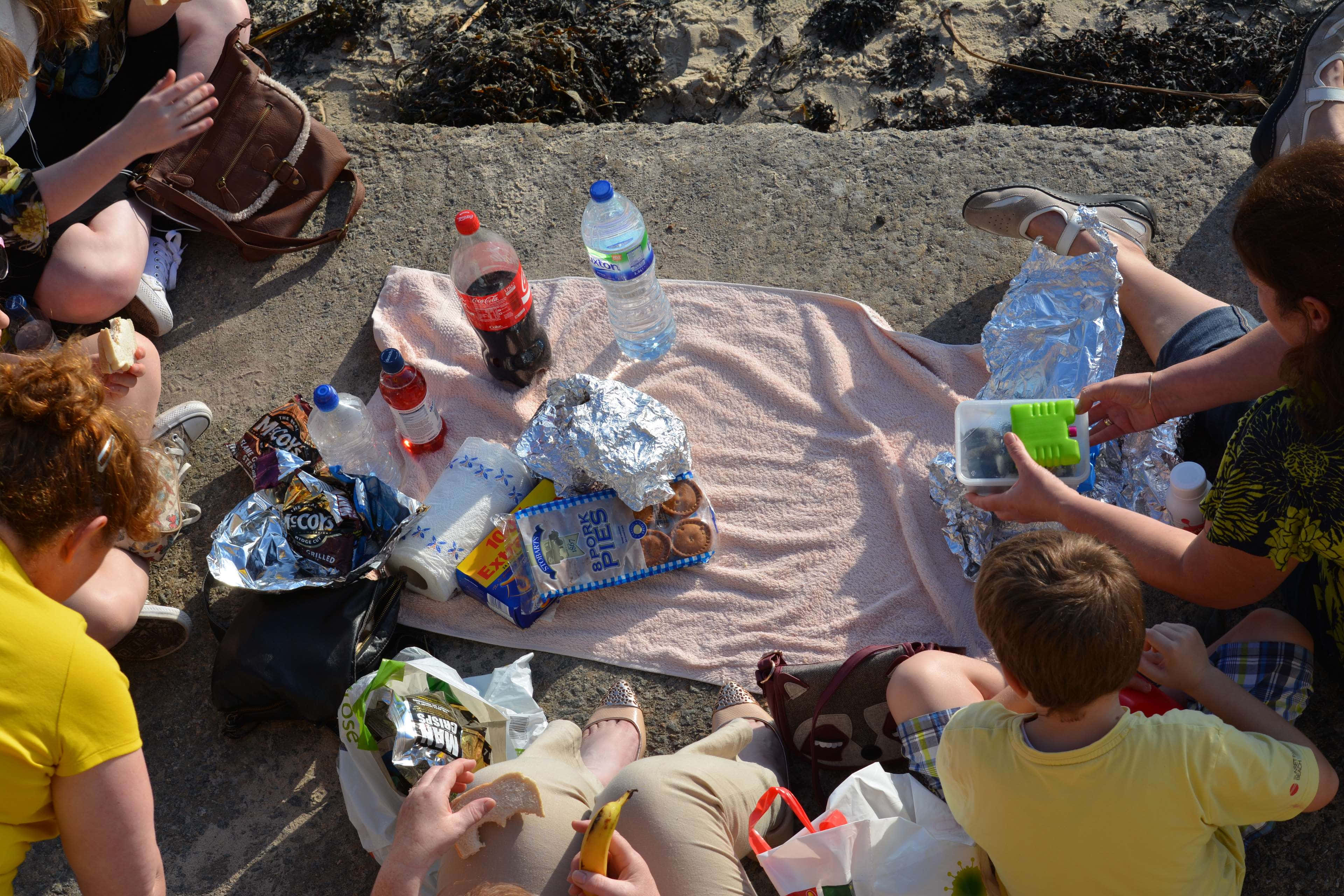 Beach Picnic 桌布 壁紙 — 一群人和小孩在粉紅色毛巾上享受陽光沙灘野餐，有零食、飲料和錫紙包裹的食物。