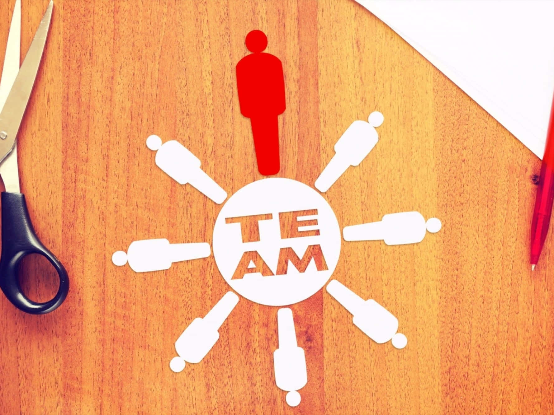 一群人在圓圈中站立 壁紙 — 一個紅色紙剪出的人站在圓形「TEAM」標誌上，周圍有白色人物，木桌上附近有剪刀和筆。