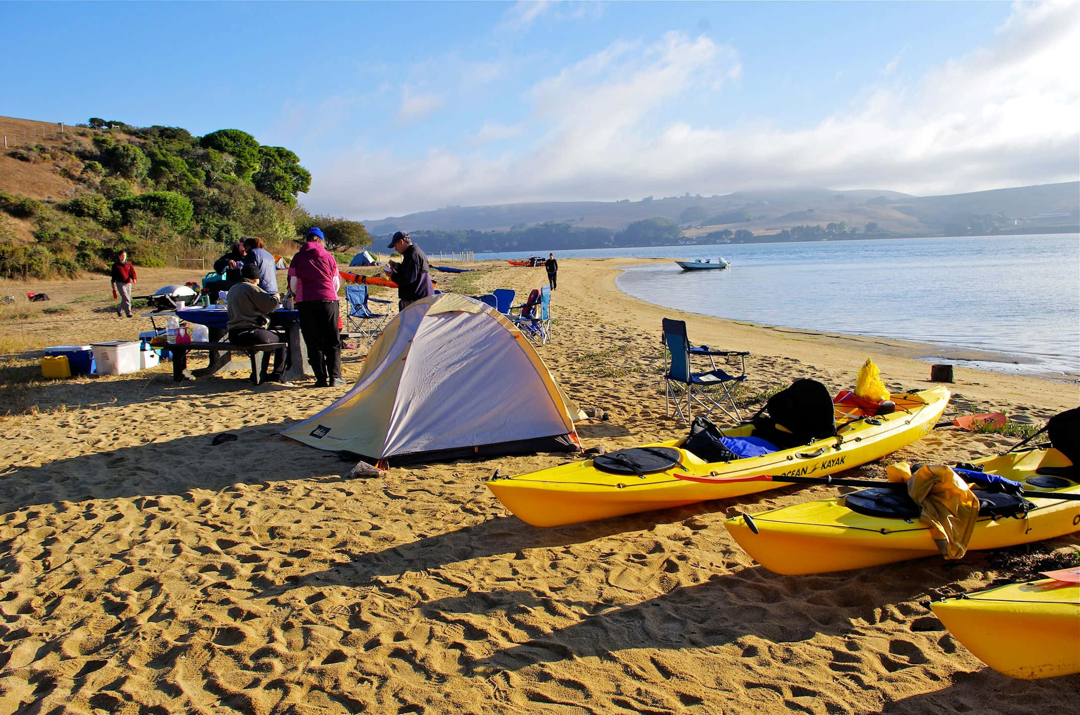 Beach Camping 桌布 壁紙 — 一群人在陽光明媚的沙灘上露營，附近有平靜的水面和起伏的山丘，還有黃色皮划艇和白色帳篷。