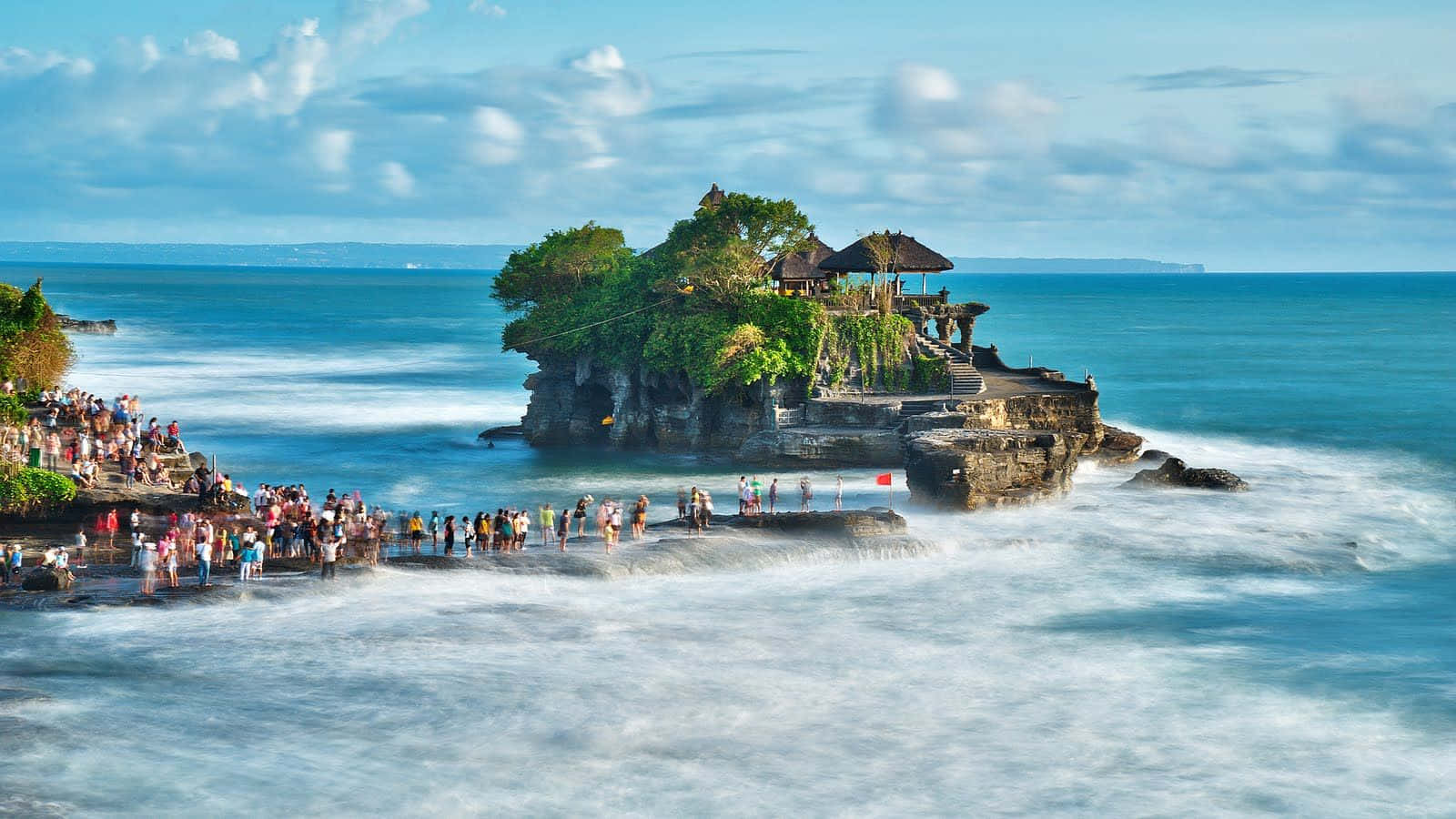 Bali Beach 桌布 壁紙 — 一群充滿活力的人群聚集在岩石島上的海神廟，碧綠的海浪在晴朗的藍天和白雲下拍打。