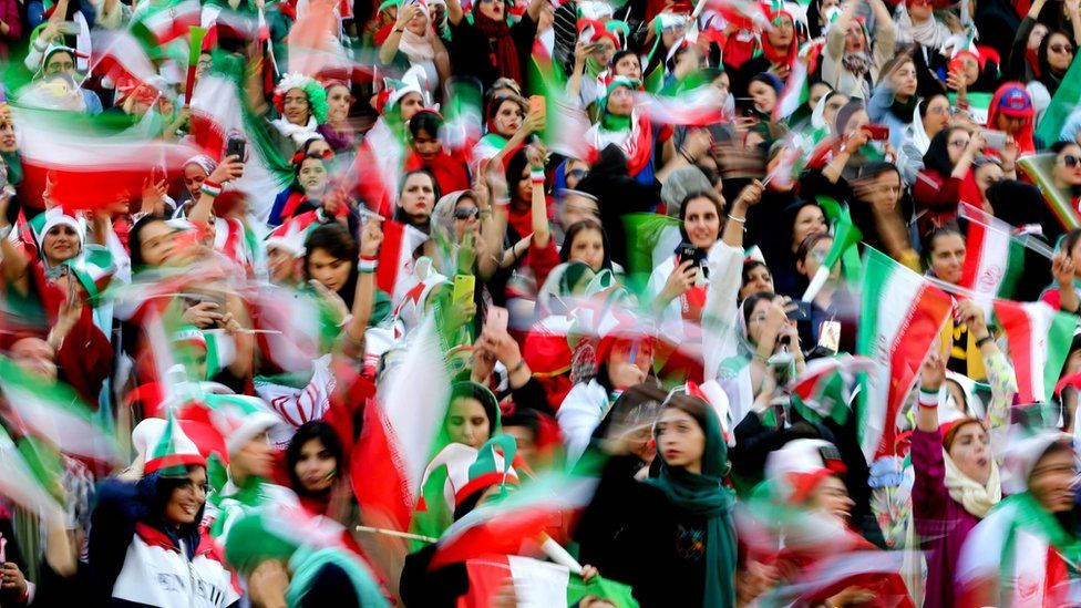 Iran 桌布 壁紙 — 一群充滿活力的伊朗球迷揮舞著紅、白、綠旗，運動模糊捕捉了體育場活動中的熱情慶祝。
