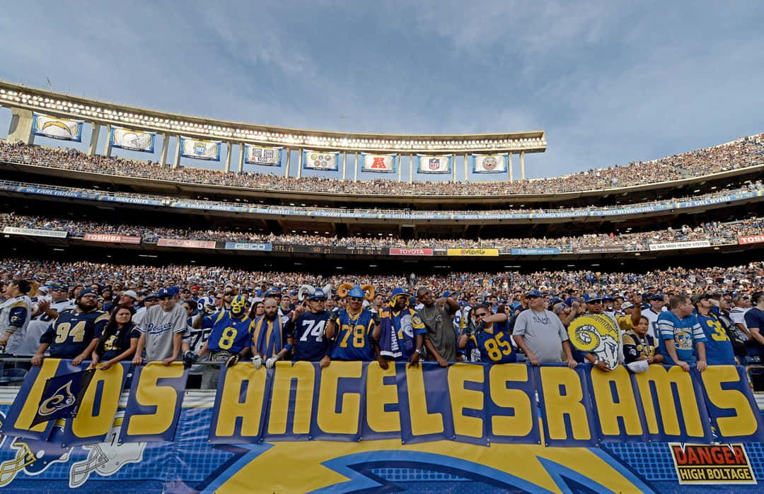 Rams 桌布 壁紙 — 一群充滿活力的洛杉磯公羊隊球迷身穿藍色和黃色球衣，在晴朗的天空下於座無虛席的體育場內歡呼。