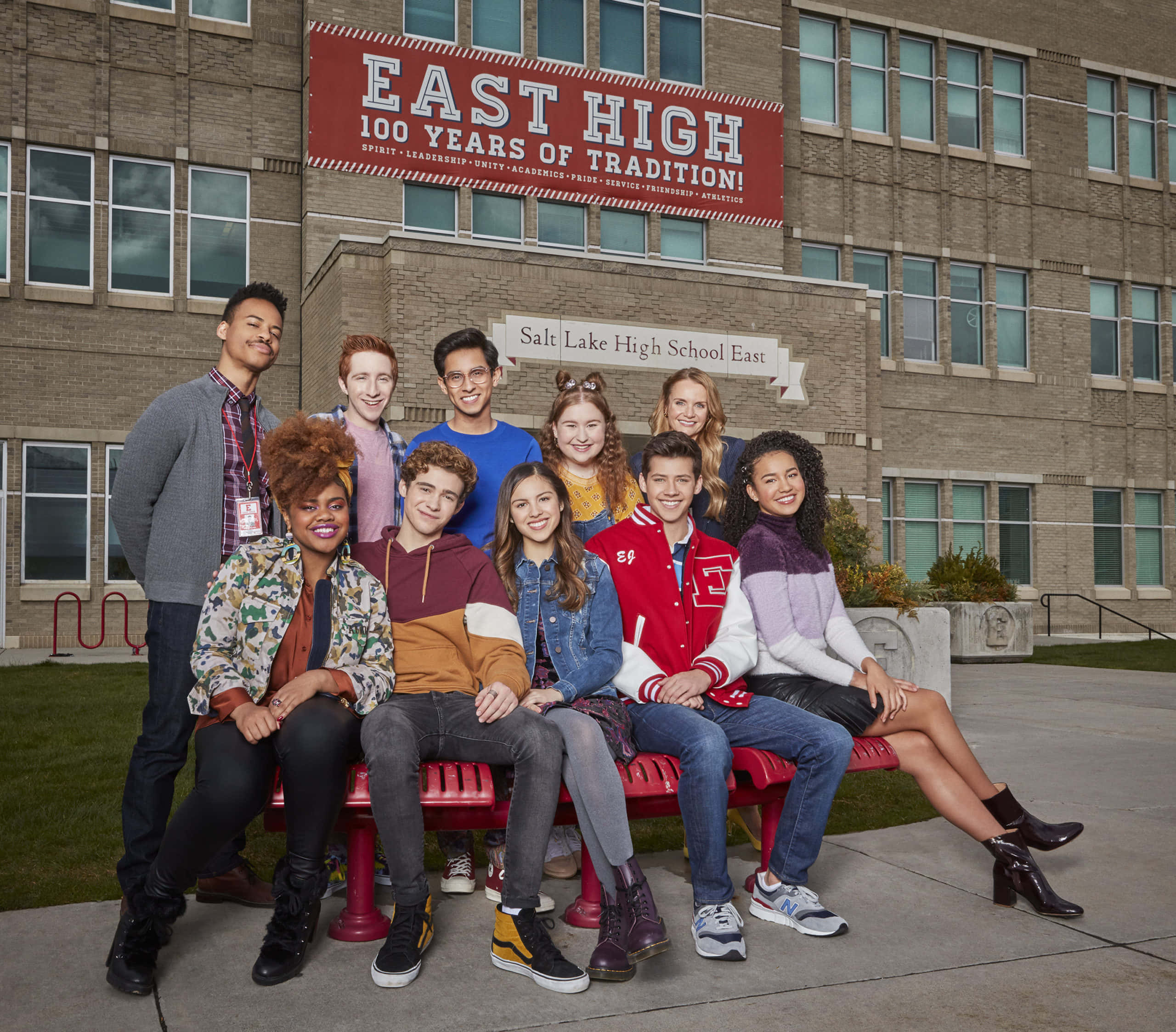 High School Musical 桌布 壁紙 — 一群多元化的高中學生在明亮的日光下，於東高中磚砌外牆前的紅色長椅上愉快地擺姿勢。