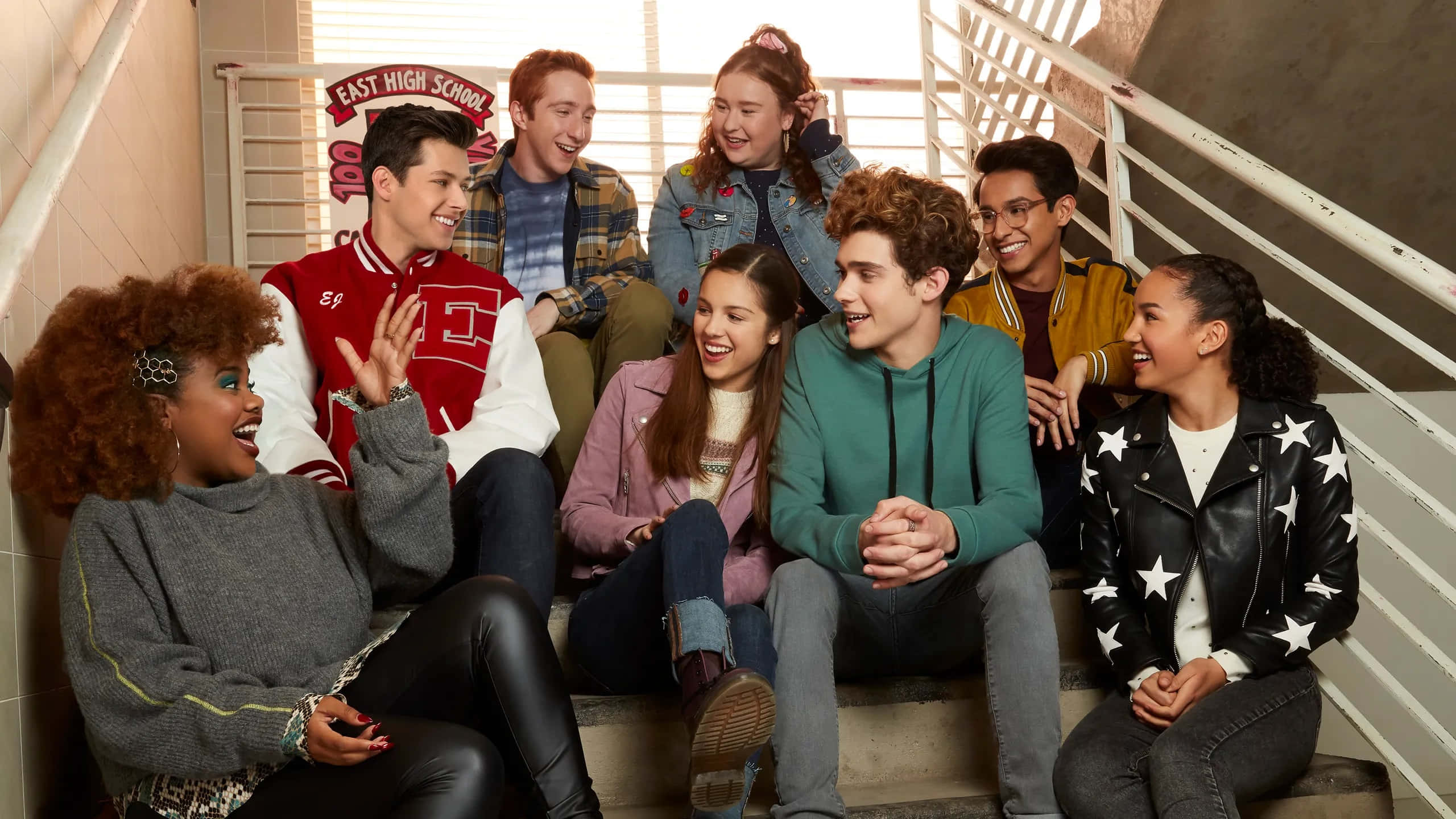 High School Musical 桌布 壁紙 — 一群多元化的高中學生坐在 'East High School' 標誌附近的混凝土樓梯上，笑著聊天。