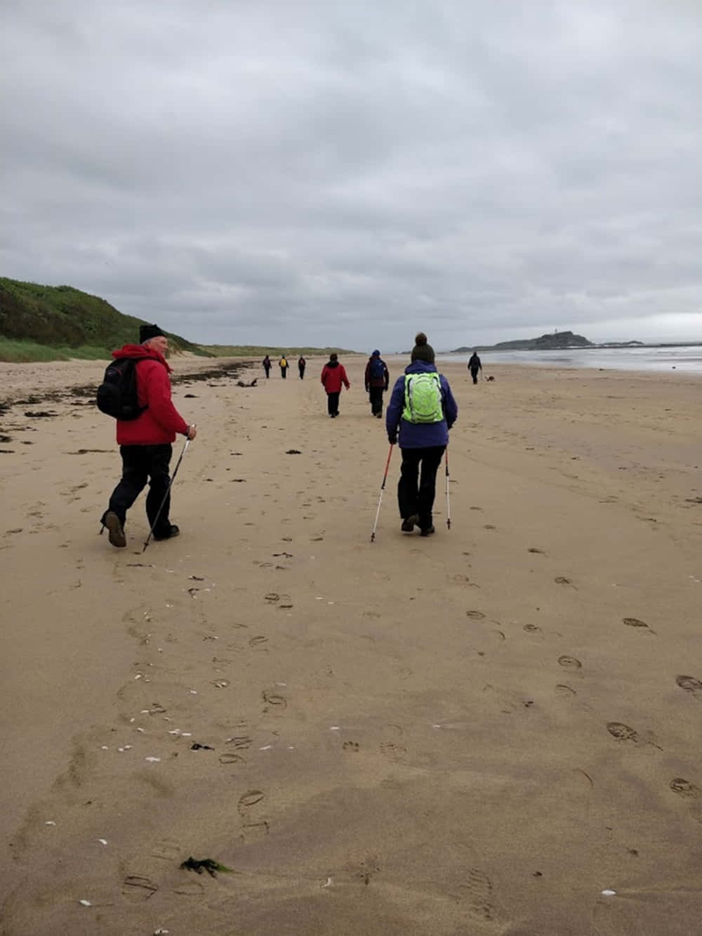 Beach Walk 桌布 壁紙 — 一群拿著登山杖的遠足者，在陰天、遠方有懸崖和波浪的濕沙灘上行走。