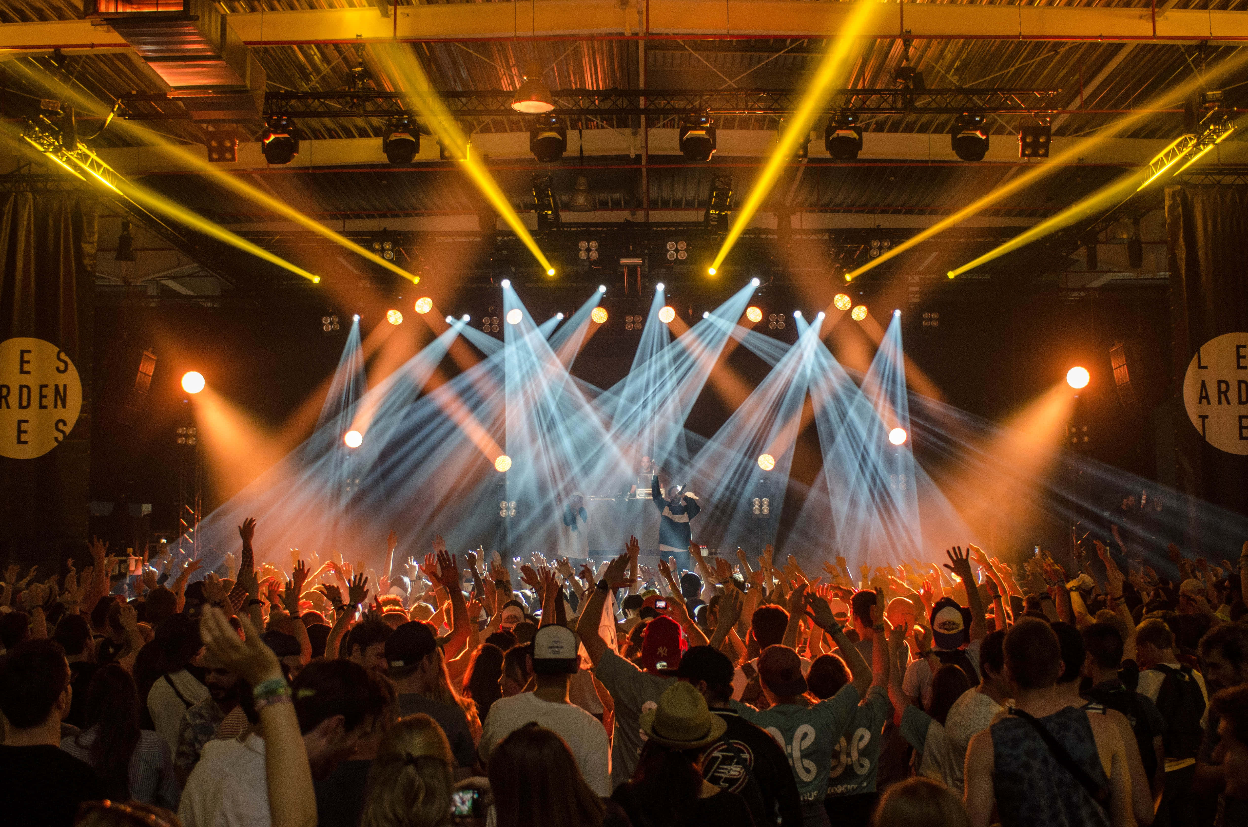 music festival 桌布 壁紙 — 一群演唱會觀眾在工業風場地中，於鮮黃色和藍色的舞台燈光下舉起雙手。