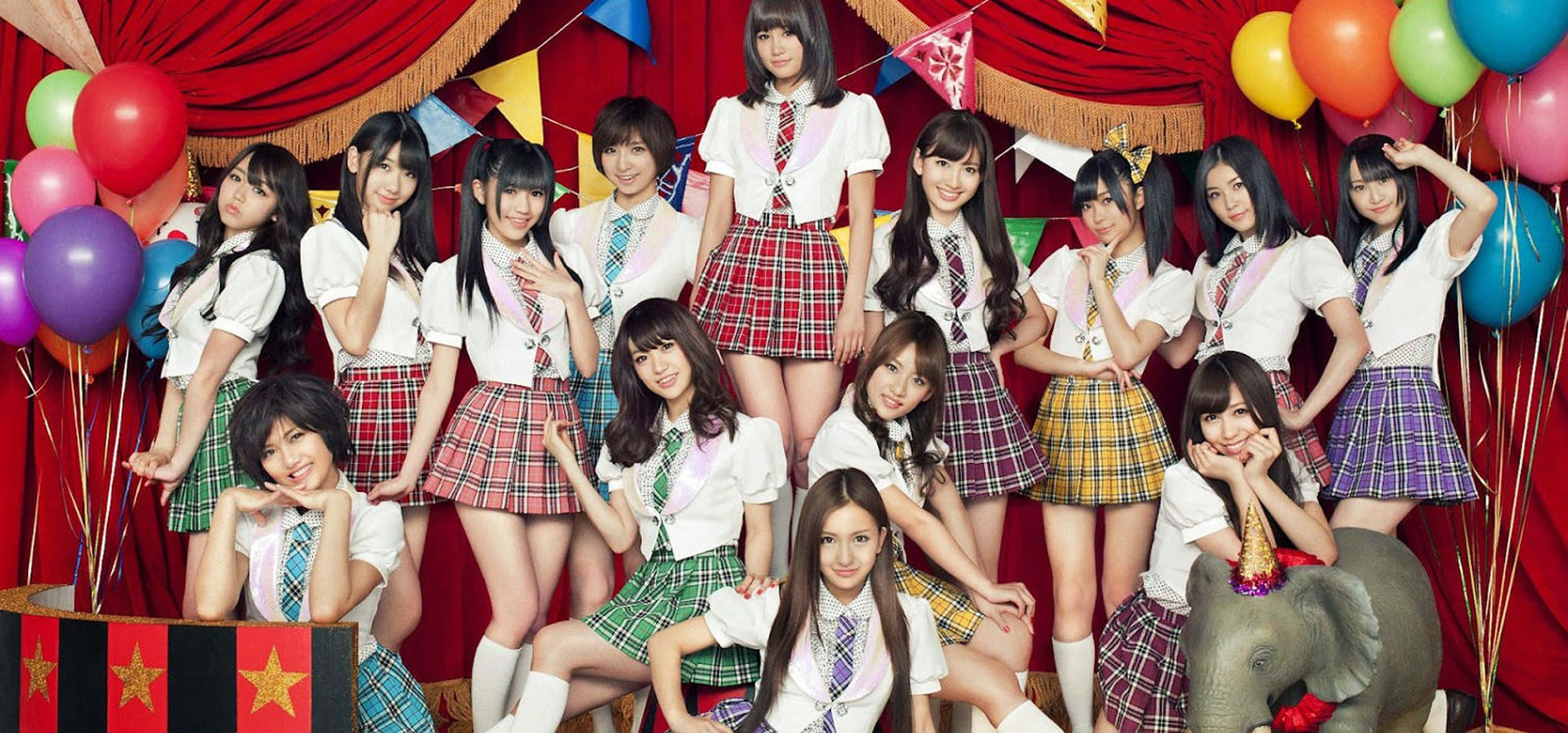 J-pop 桌布 壁紙 — 一群穿著彩色格子裙和白色襯衫的年輕女性，在裝飾著氣球和派對旗幟的紅色窗簾背景前愉快地擺姿勢。