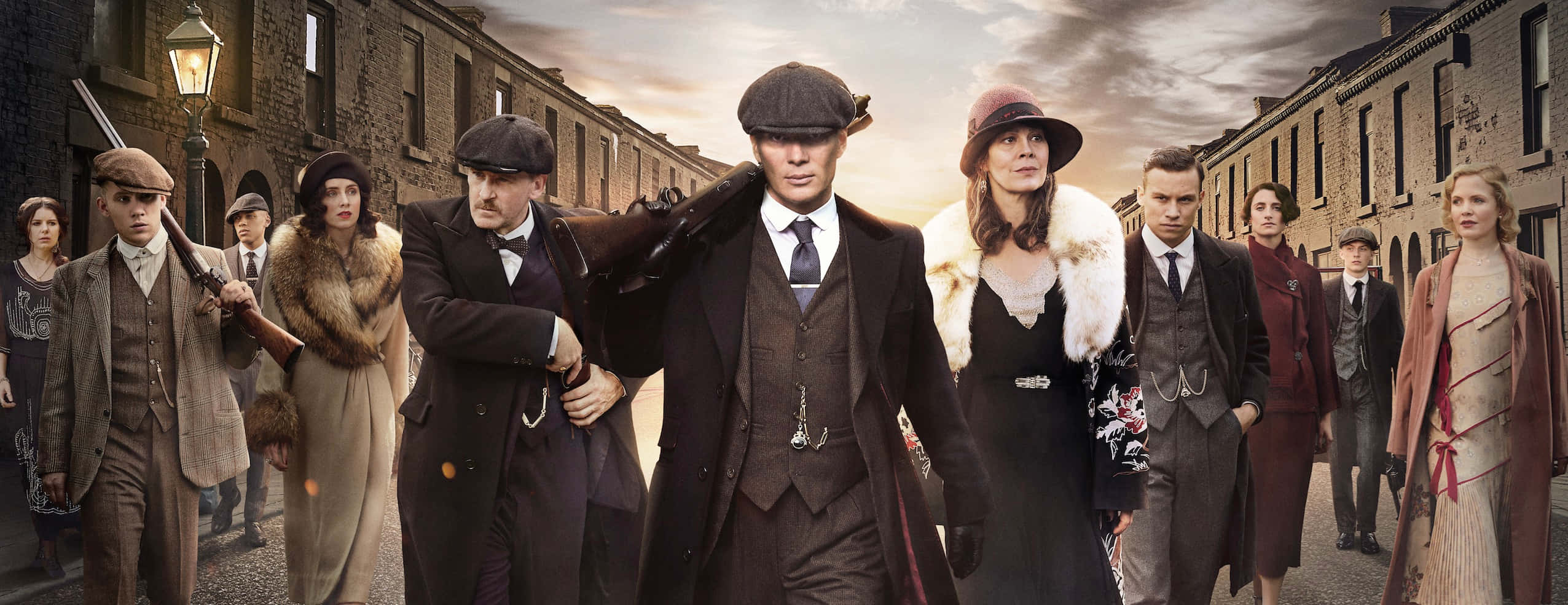 Peaky Blinders 桌布 壁紙 — 一群穿著復古大衣和帽子的時代角色，在陰天中沿著狹窄的磚街大步前行。