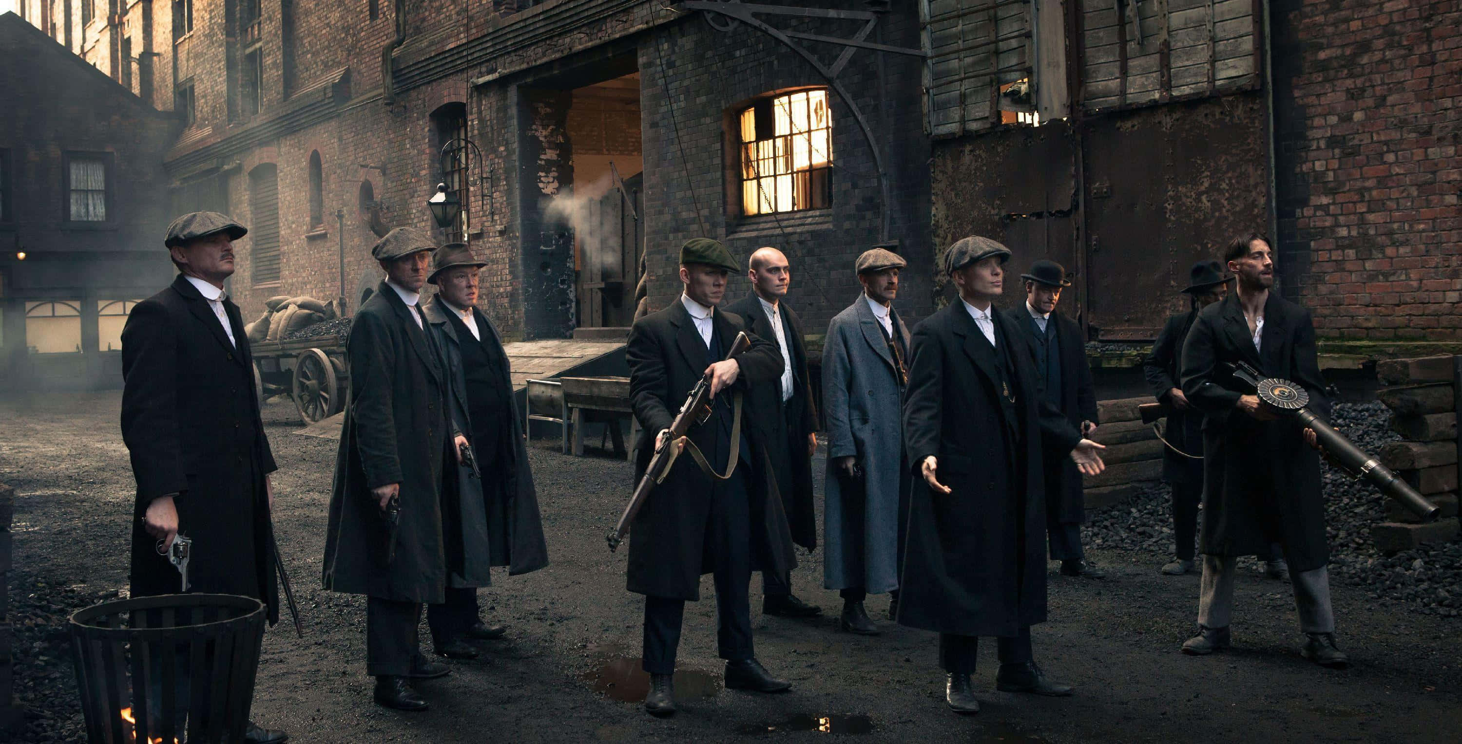 Peaky Blinders 桌布 壁紙 — 一群穿著時代外套和帽子的男人，在昏暗的工廠燈光下，手持武器站在骯髒的工業小巷中。