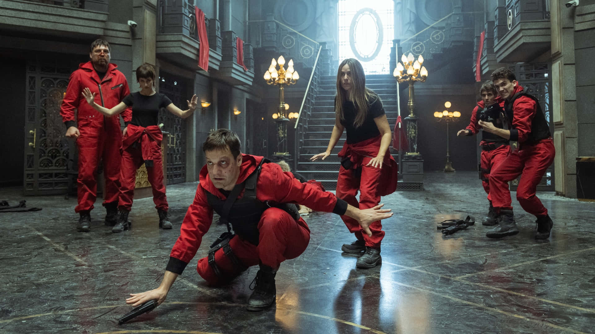 Money Heist 桌布 壁紙 — 一群穿著紅色連身衣褲的人緊張地蹲下和警覺地站立在一個宏偉、燈光昏暗的大宅走廊中，走廊有華麗的水晶吊燈和大理石地板。