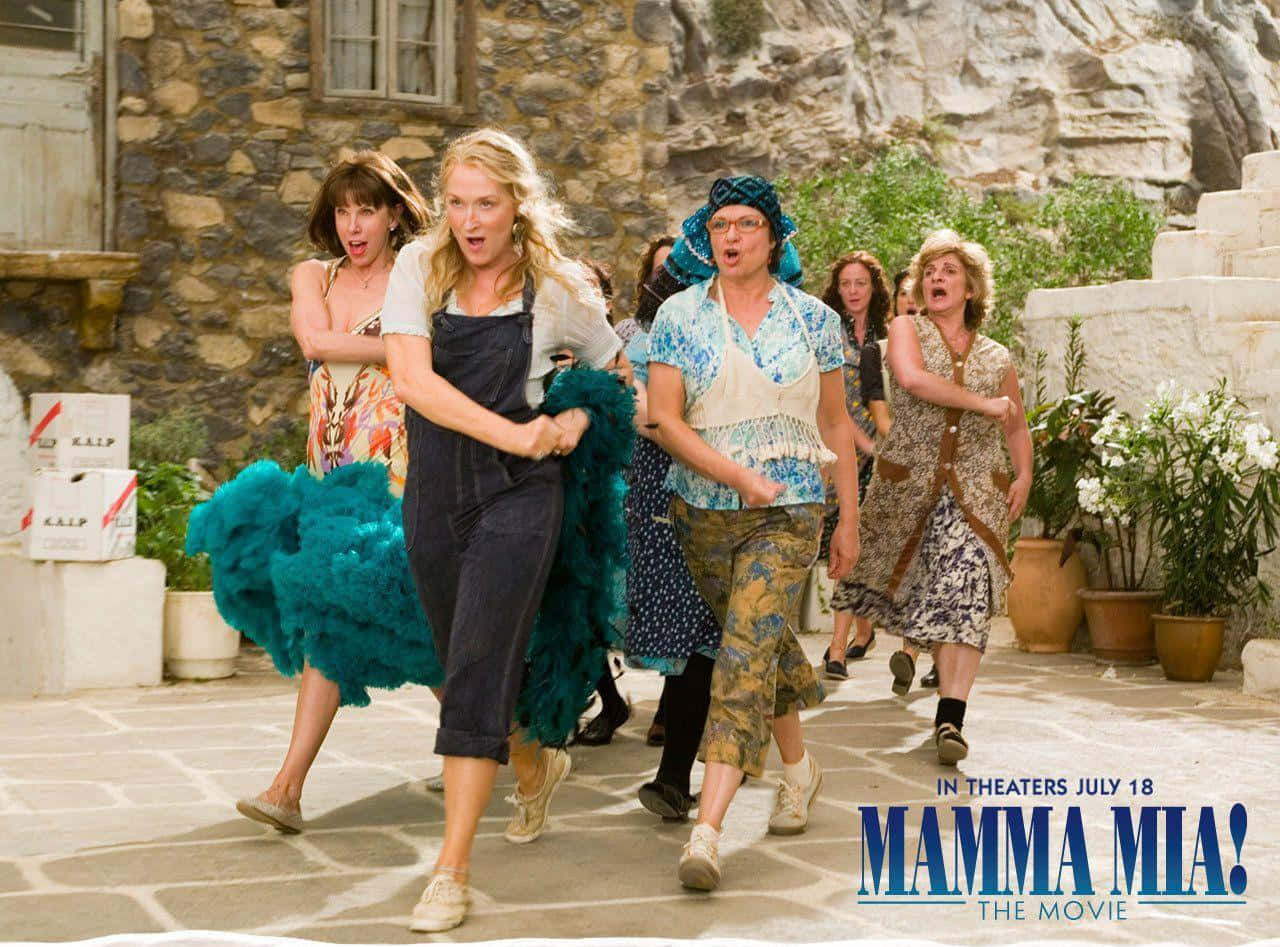 Mamma Mia 桌布 壁紙 — 一群穿著色彩繽紛服裝的女性，在陽光普照的石頭庭院中快樂地奔跑，背景是岩石峭壁。