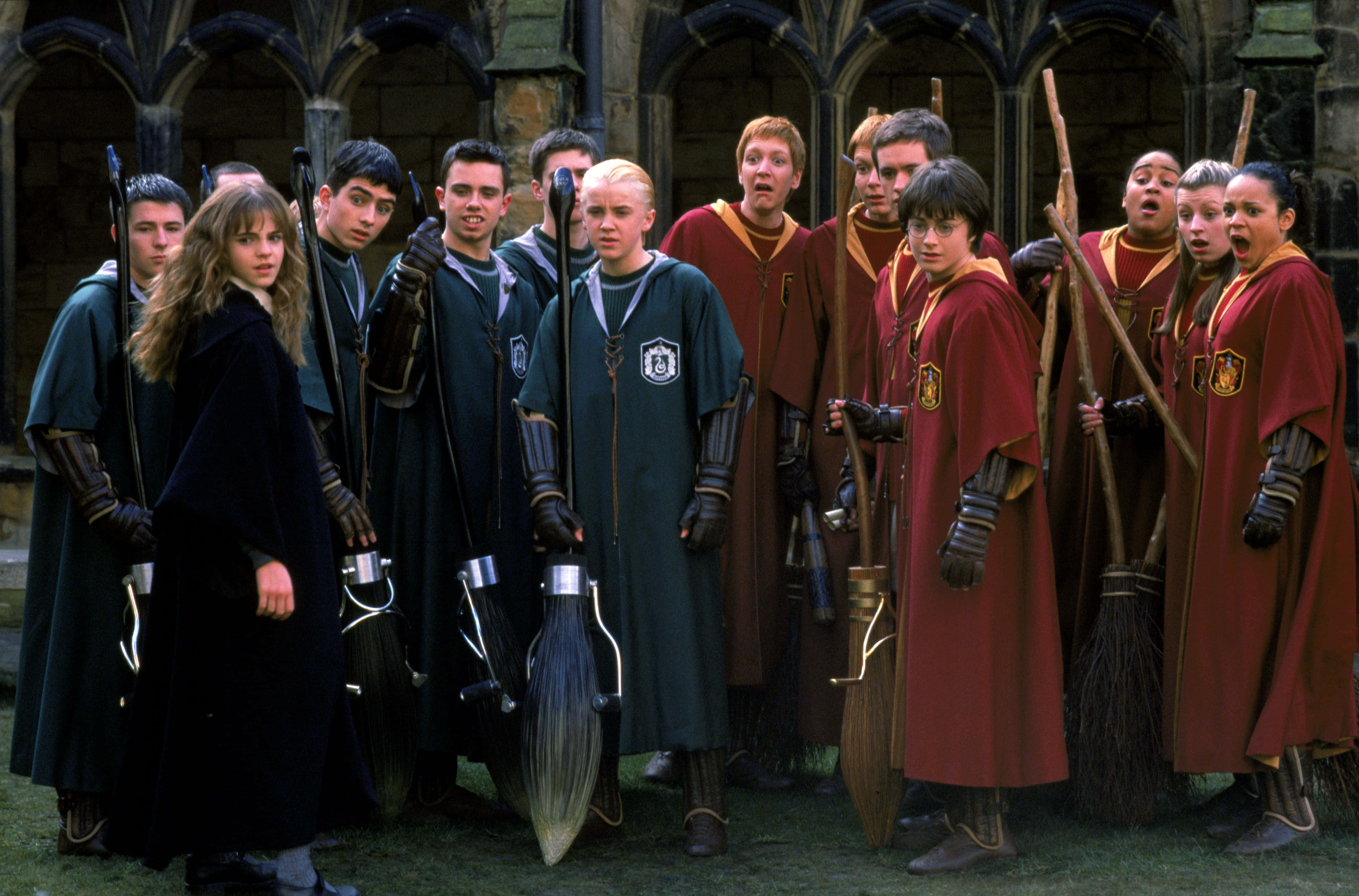 The Hogwarts Quidditch Team 桌布 壁紙 — 一群穿著霍格華茲制服的年輕女巫和巫師站在石頭庭院中，手持掃帚，表情驚訝。