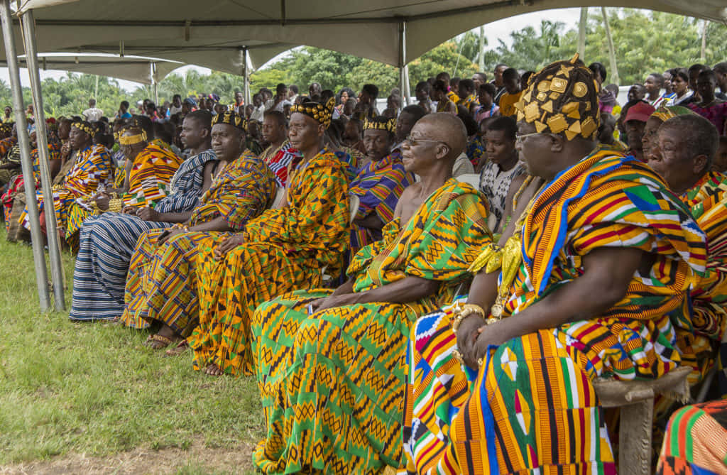 African Culture 桌布 壁紙 — 一群穿著鮮豔傳統非洲服飾的人群，在草地戶外環境中，專注地坐在白色帳篷下。