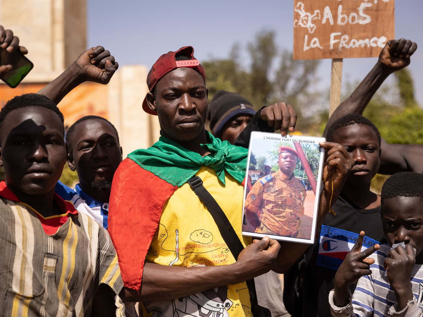 African 桌布 壁紙 — 一群穿著鮮豔衣服的抗議者在晴朗的白天示威期間舉起一名士兵的裱框照片和標語牌。