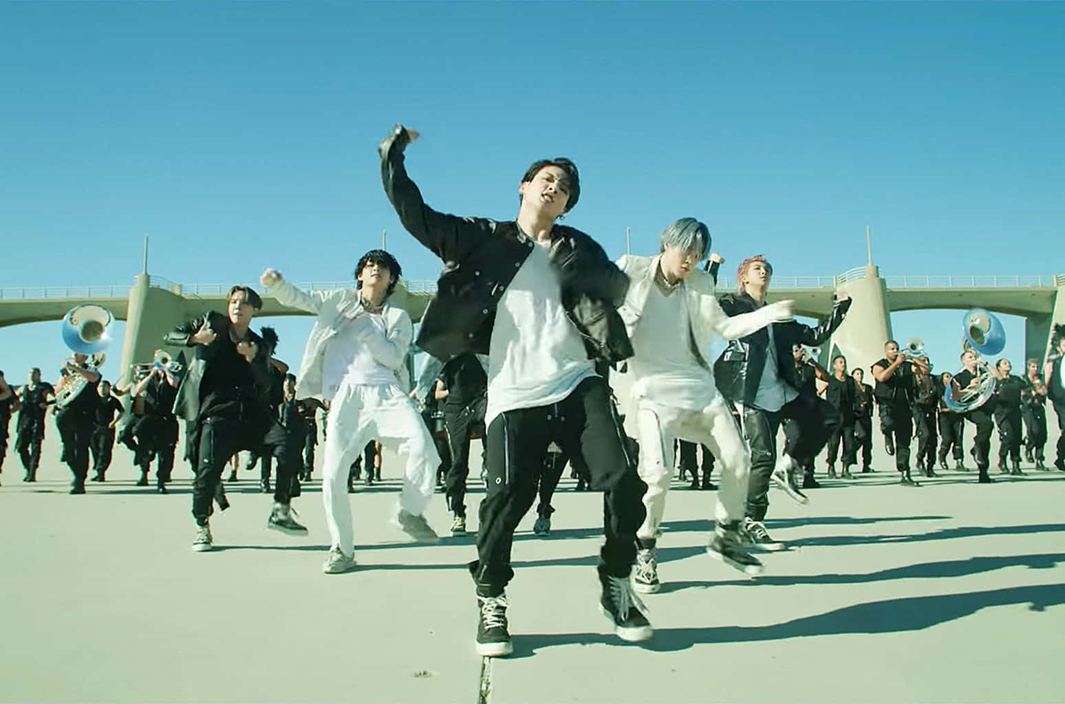 Bts Mv 桌布 壁紙 — 一群穿著黑白服裝的年輕男子在晴朗的藍天下充滿活力地跳舞，背景有橋樑和遊行樂隊。