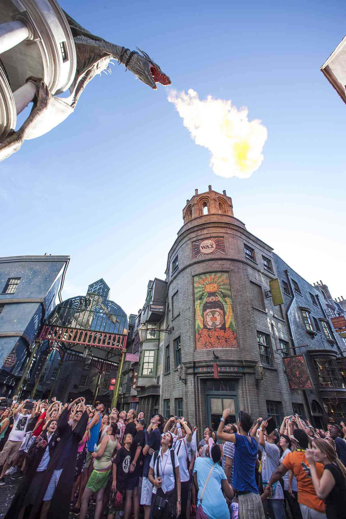 Wizarding World 桌布 壁紙 — 一群興奮的遊客觀看一條龍在晴朗藍天下於巫師街上噴火。
