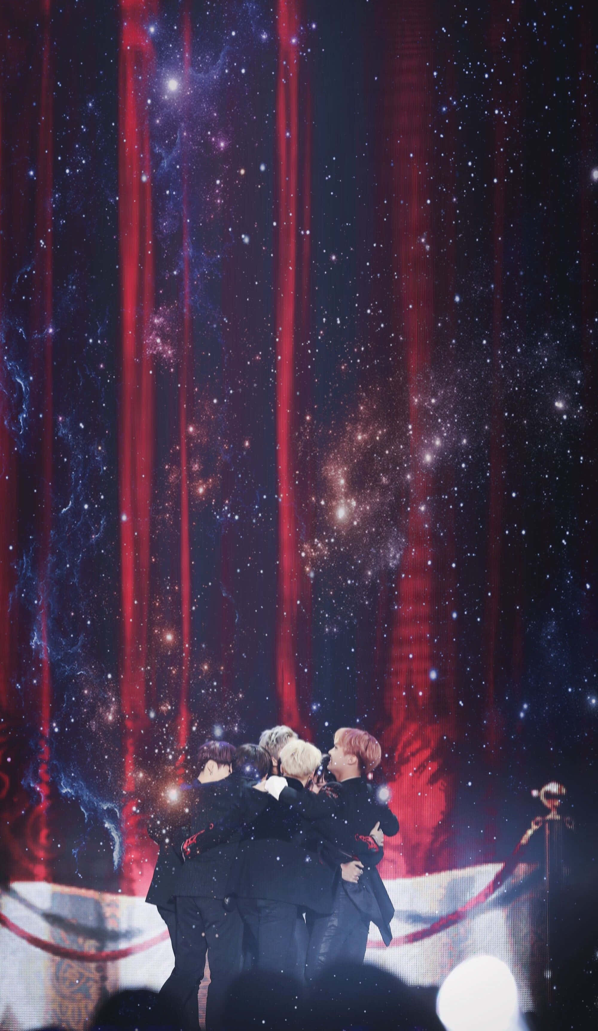 Bts Stage 桌布 壁紙 — 一群表演者在紅色燈光和閃爍星星的宇宙背景下於舞台上擁抱。