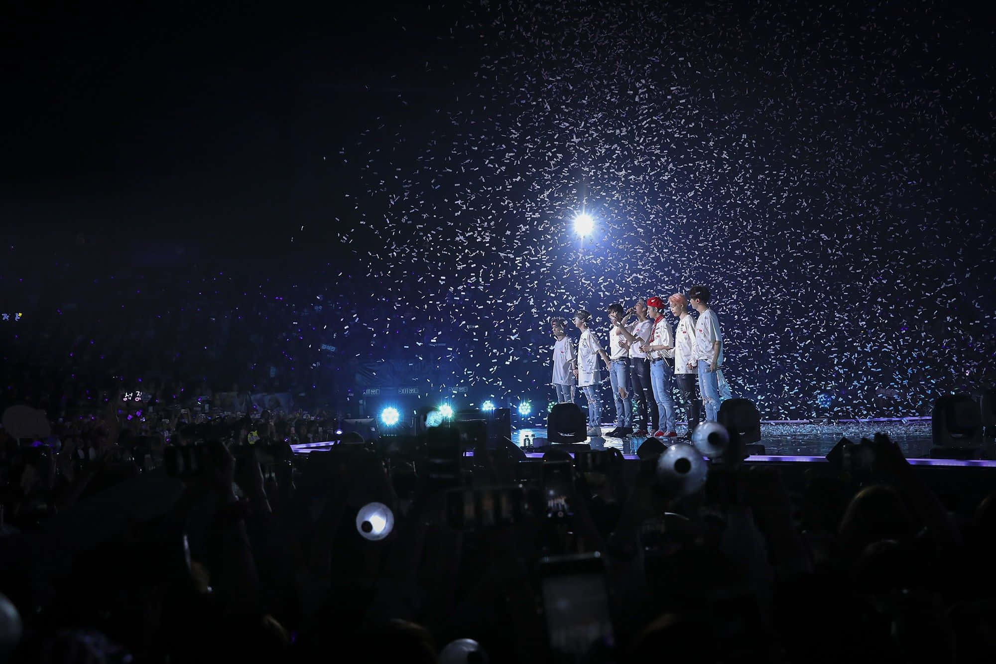 Bts Stage 桌布 壁紙 — 一群表演者夜晚站在燈光明亮的舞台上，五彩紙屑如雨般灑落在下方黑暗的人群中。