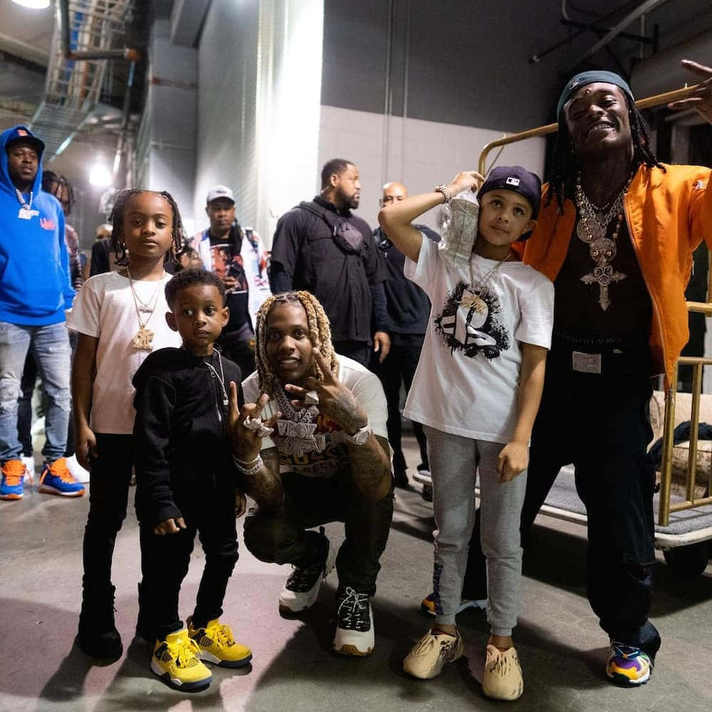 lil durk 桌布 壁紙 — 一群說唱歌手和兒童在昏暗的後台區域，在工業照明和金屬結構下一起擺姿勢。