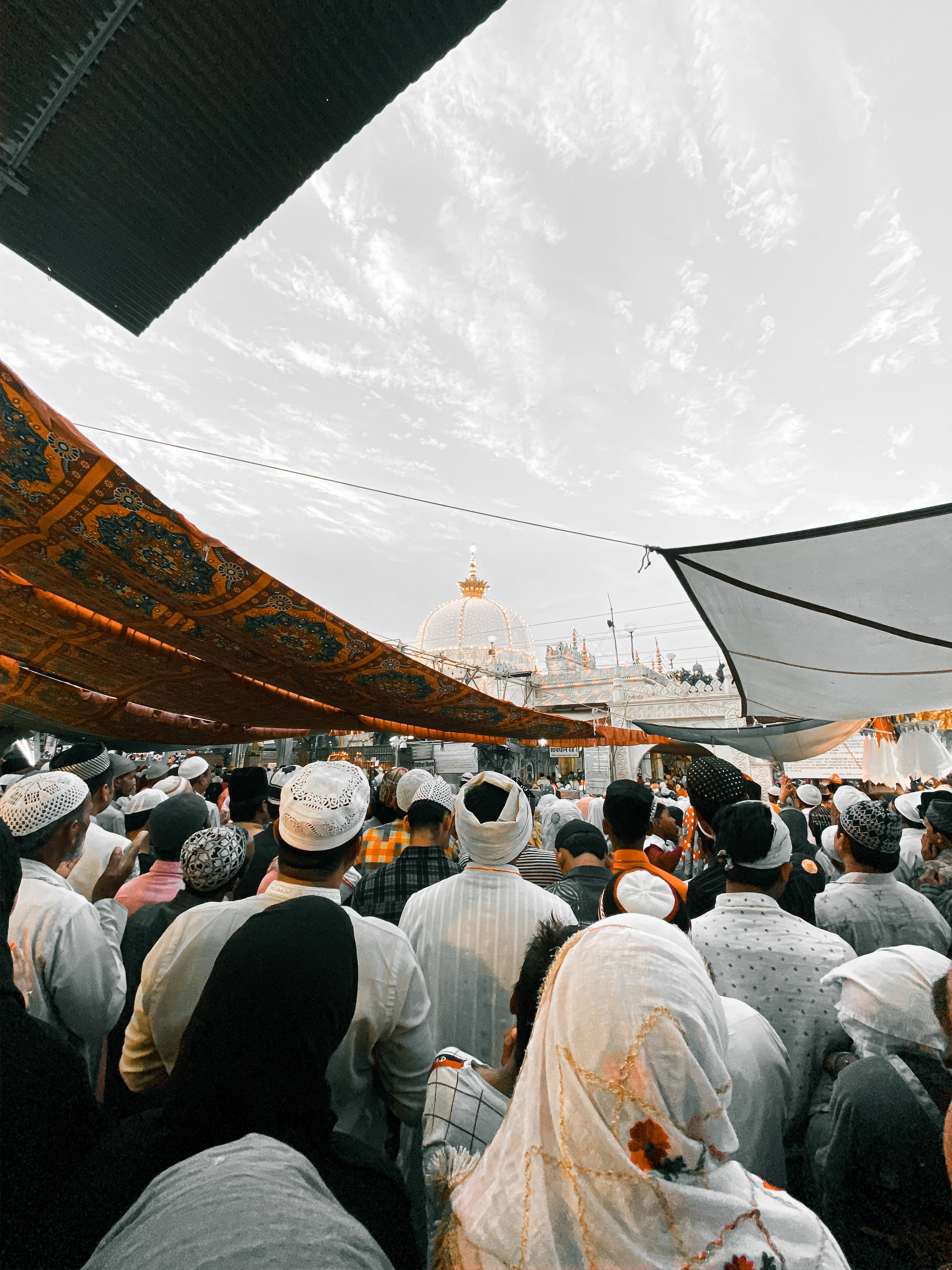 muslim 桌布 壁紙 — 一群身穿傳統服飾的朝拜者在有圖案的遮篷下聚集，朝向遠處一座金色圓頂聖殿，天空多雲。