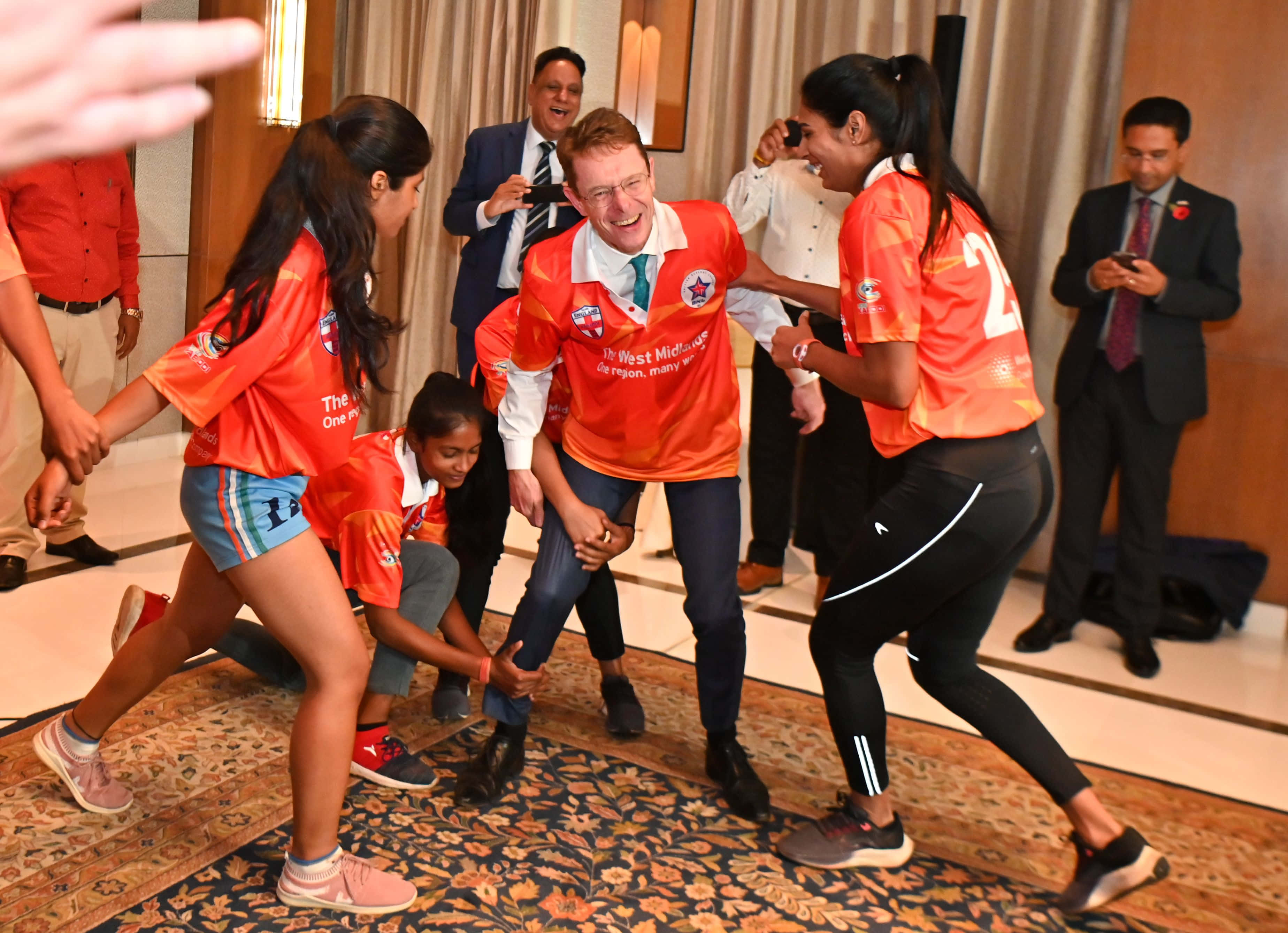 Kabaddi 桌布 壁紙 — 一群身穿橙色球衣的年輕運動員在熱鬧的室內活動中，頑皮地擒抱一位微笑的西裝男子。