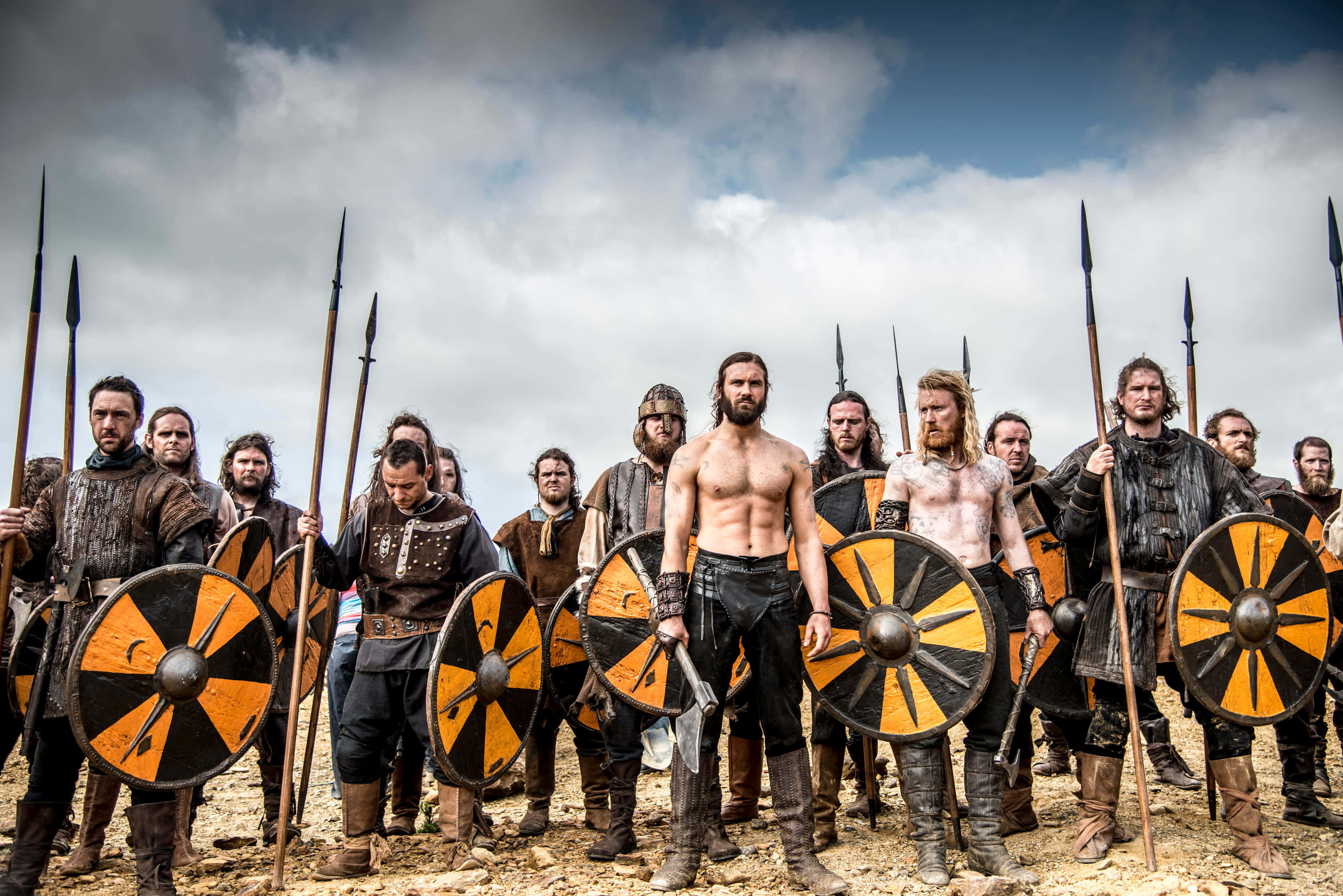 Vikings 桌布 壁紙 — 一群身穿皮甲、手持橙黑色盾牌的維京戰士在陰天準備就緒。