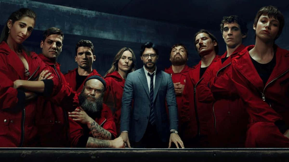 Money Heist 桌布 壁紙 — 一群身穿紅色連身衣褲的強烈角色和一名身穿西裝的男子站在光線昏暗的工業空間中。