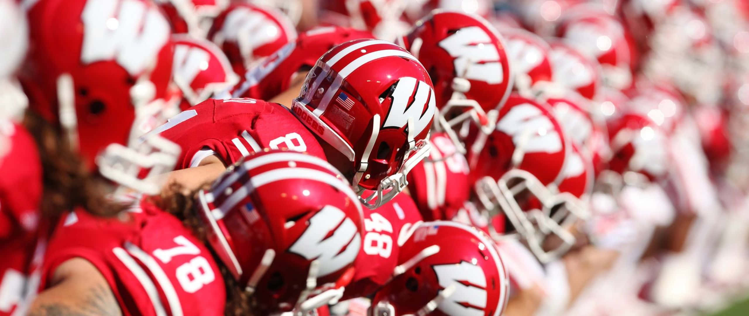 Wisconsin Badgers 桌布 壁紙 — 一群身穿紅色頭盔和球衣的美式足球員在陽光明媚的球場上緊密地聚在一起。