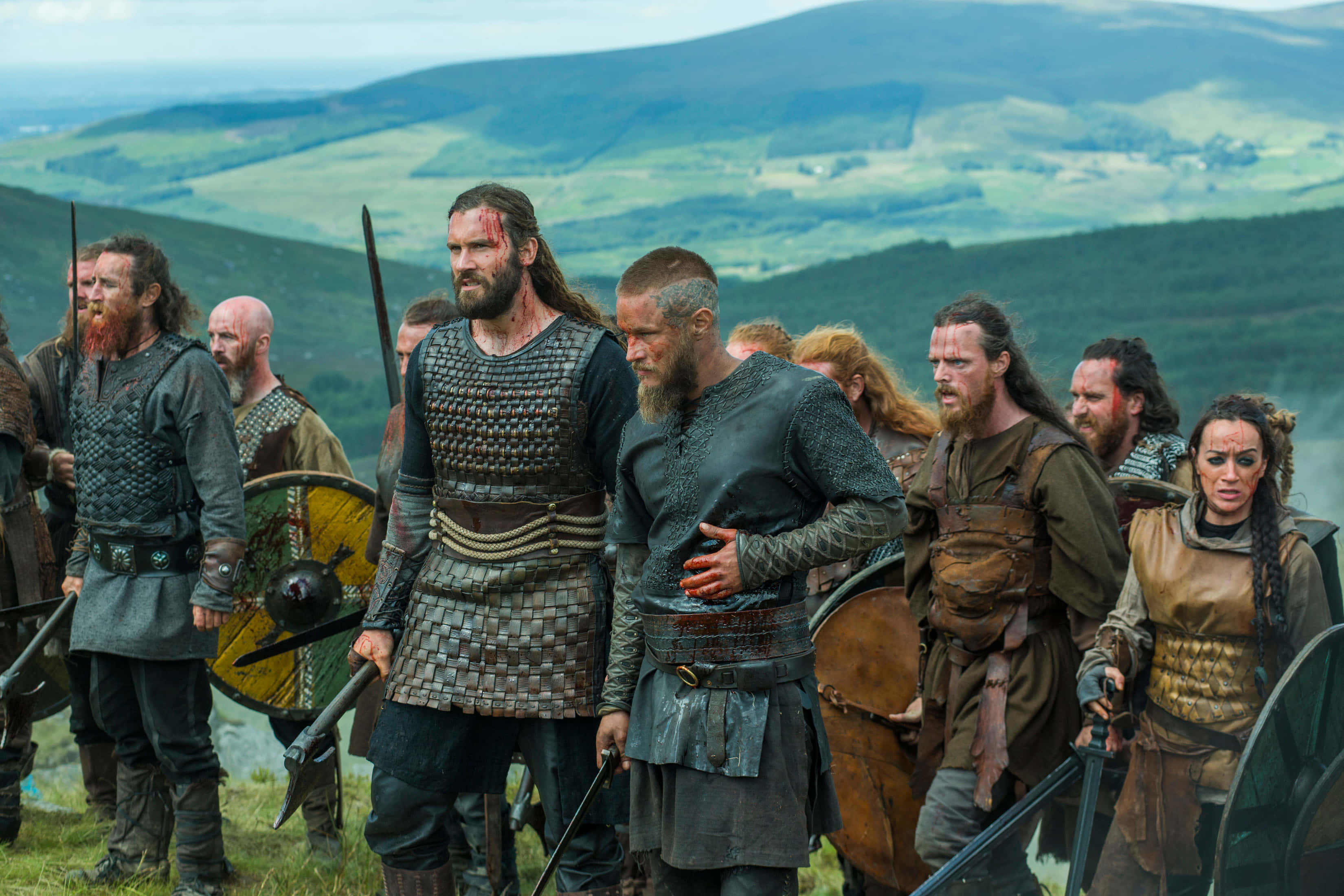 Vikings 桌布 壁紙 — 一群身穿鎖子甲和皮革盔甲的維京戰士莊嚴地站在一個草坡上，俯瞰著連綿起伏的綠色山丘。