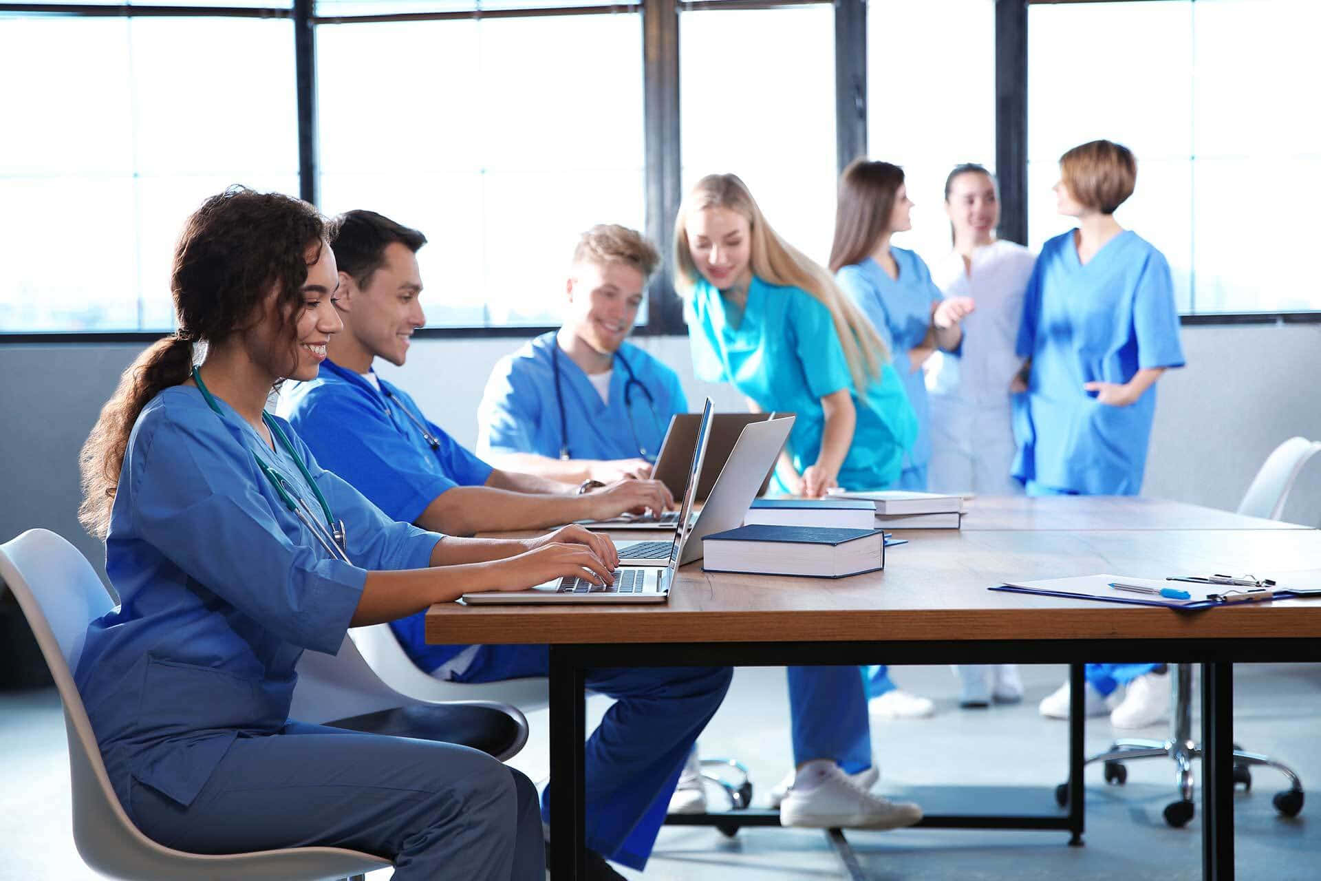 Nursing 桌布 壁紙 — 一群面帶微笑的醫護專業人員穿著藍色手術衣，在明亮的現代辦公室裡圍著木桌，使用手提電腦和書籍進行協作。