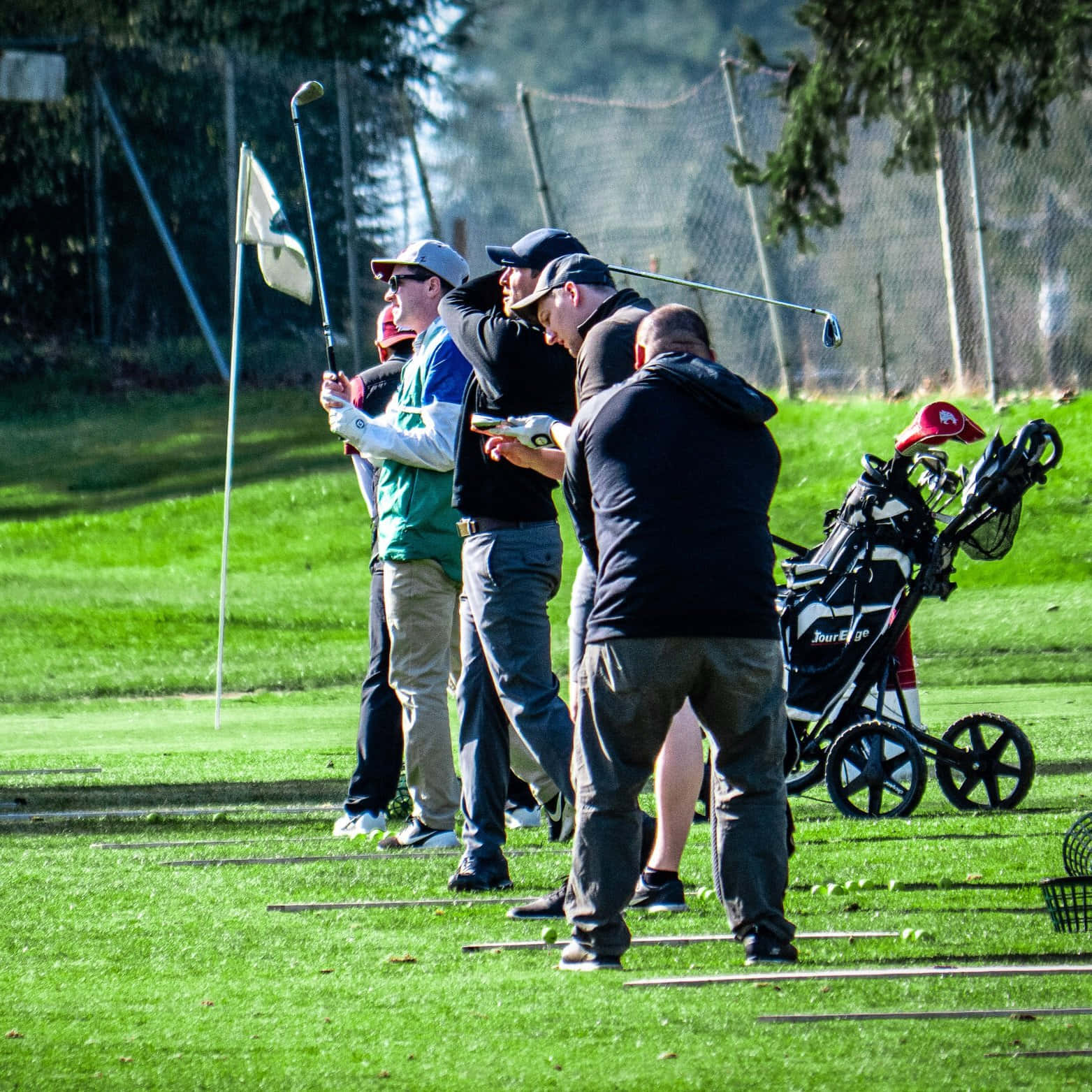 Golf Green 桌布 壁紙 — 一群高爾夫球手聚集在陽光明媚的綠色球場上，一人正在揮桿，其他人則帶著球桿和球袋在附近觀看。