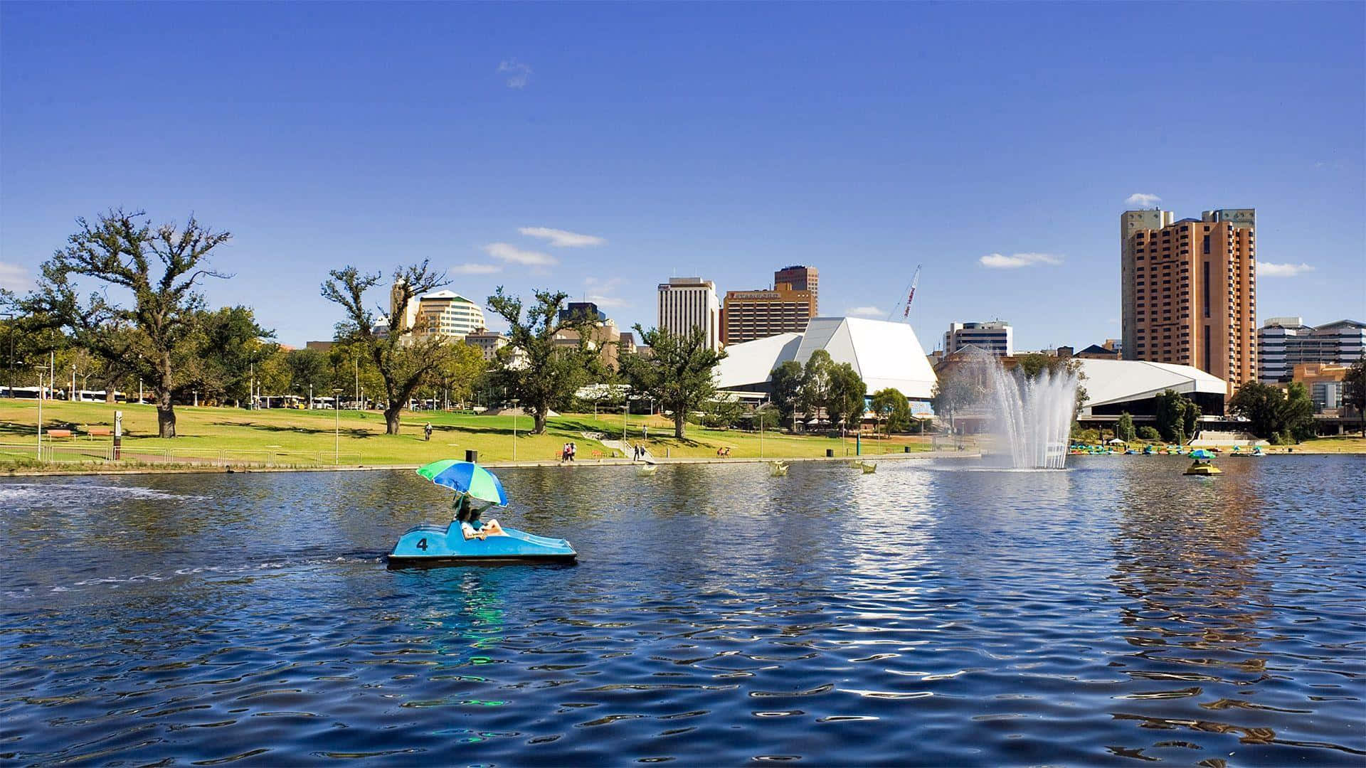 Adelaide 桌布 壁紙 — 一艘亮藍色的腳踏船帶著彩色遮陽傘，在晴朗藍天下平靜的湖面上滑行，背景是城市天際線和噴泉。
