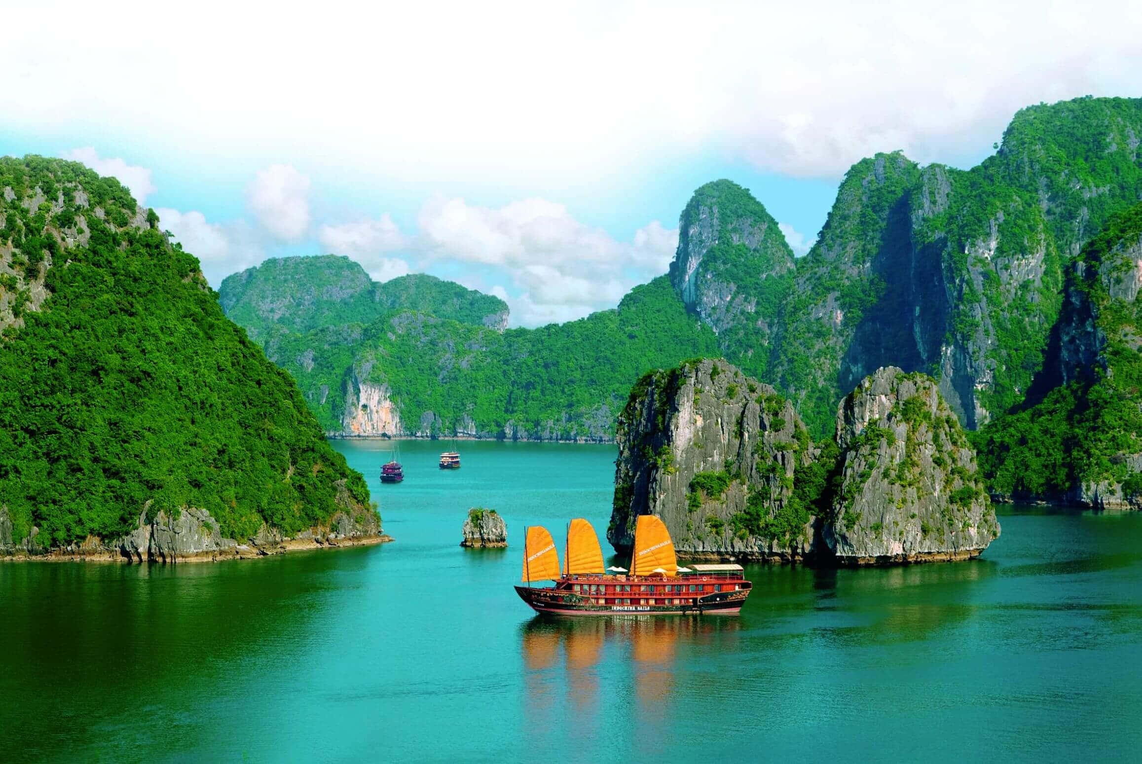 Vietnam 桌布 壁紙 — 一艘傳統越南帆船，帶有橙色帆，在部分多雲的天空下，滑行於被高聳的綠色喀斯特島嶼環繞的碧綠海水之中。