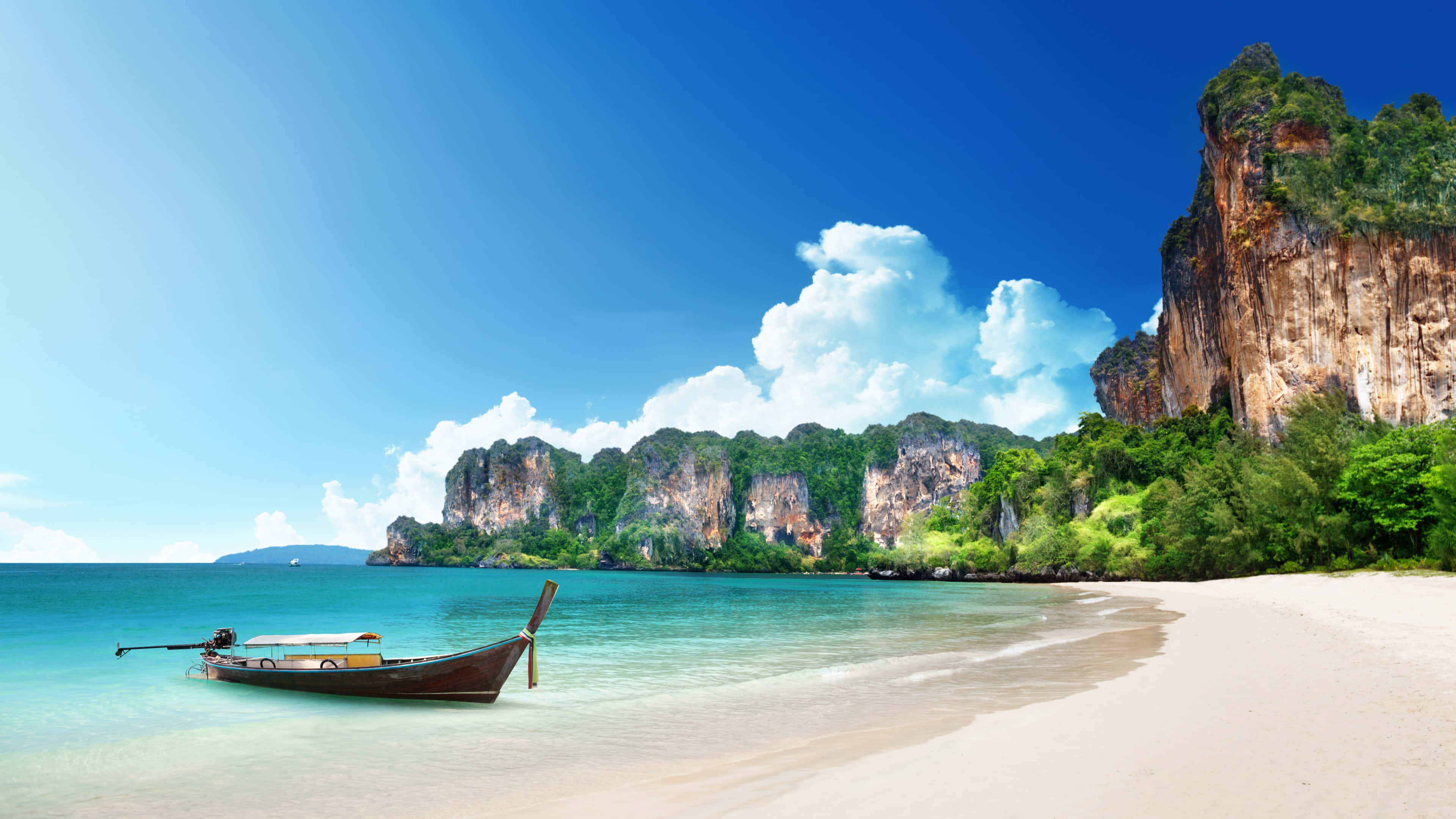 Beach Holiday 桌布 壁紙 — 一艘傳統長尾船停泊在純淨的白色沙灘上，旁邊是碧綠的海水，石灰岩懸崖在茂密的綠植後方聳立，上方是明亮的藍天。