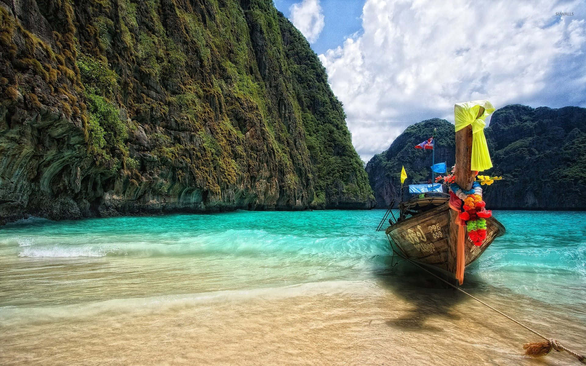 Thailand Beach 桌布 壁紙 — 一艘傳統長尾船停泊在金色沙灘上，旁邊是碧綠的海水，四周是高聳的綠色懸崖，上方是明亮的藍天。