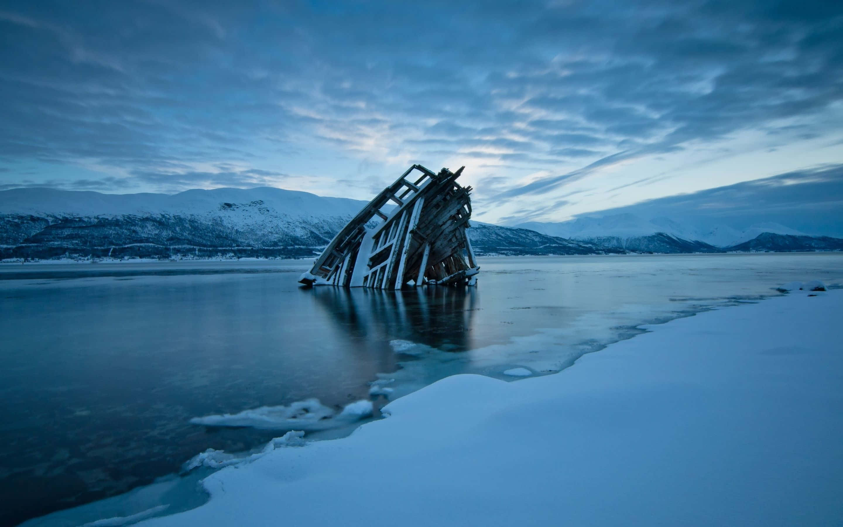 Frozen Lake 桌布 壁紙 — 一艘傾斜的沉船在冰冷的海洋中凍結，背景是陰鬱的暮色天空和遠處的雪山。