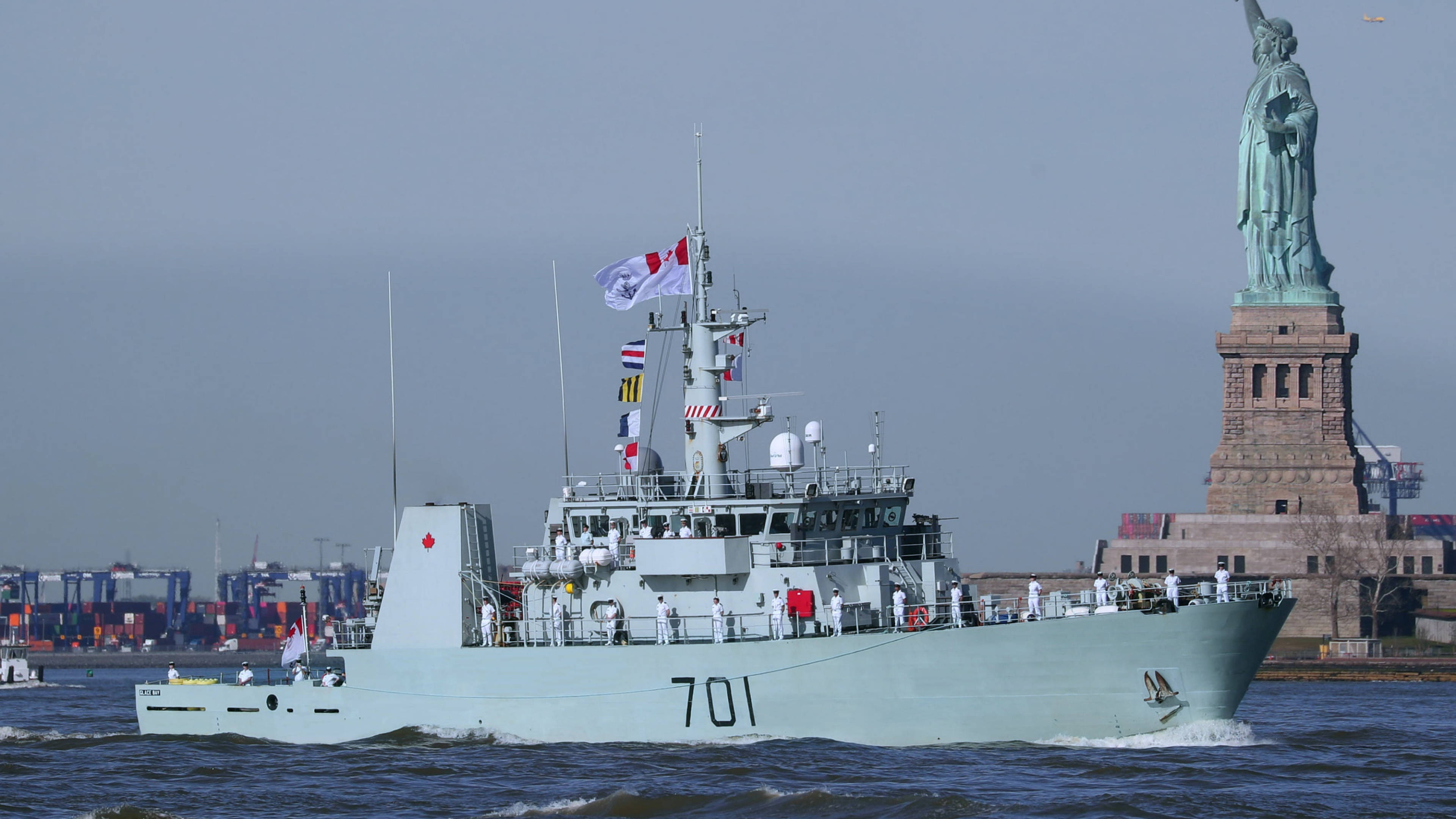 Ship 桌布 壁紙 — 一艘加拿大軍艦在多雲的天空下駛過自由女神像，遠處有城市貨櫃。