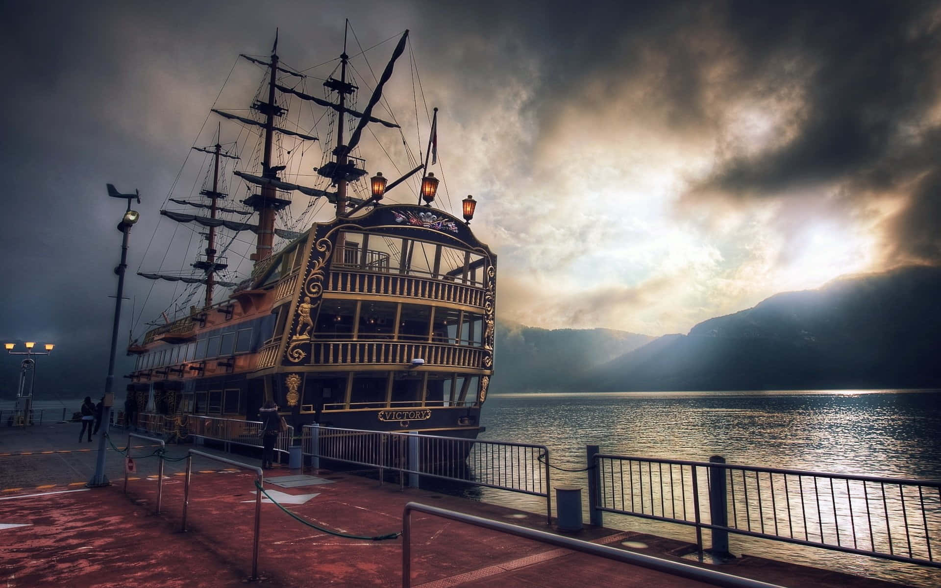 Pirate Ship 桌布 壁紙 — 一艘大型海盜船停靠在碼頭，天空戲劇性地風起雲湧，金色陽光穿透雲層。