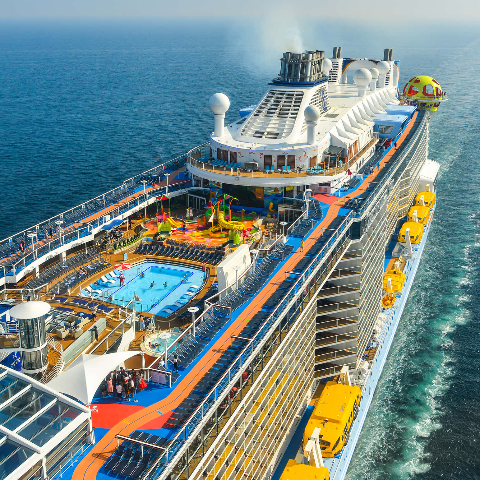 Cruise Ship 桌布 壁紙 — 一艘大型郵輪在深藍色海洋上航行的鳥瞰圖，設有色彩繽紛的泳池、水上滑梯和黃色接駁船。