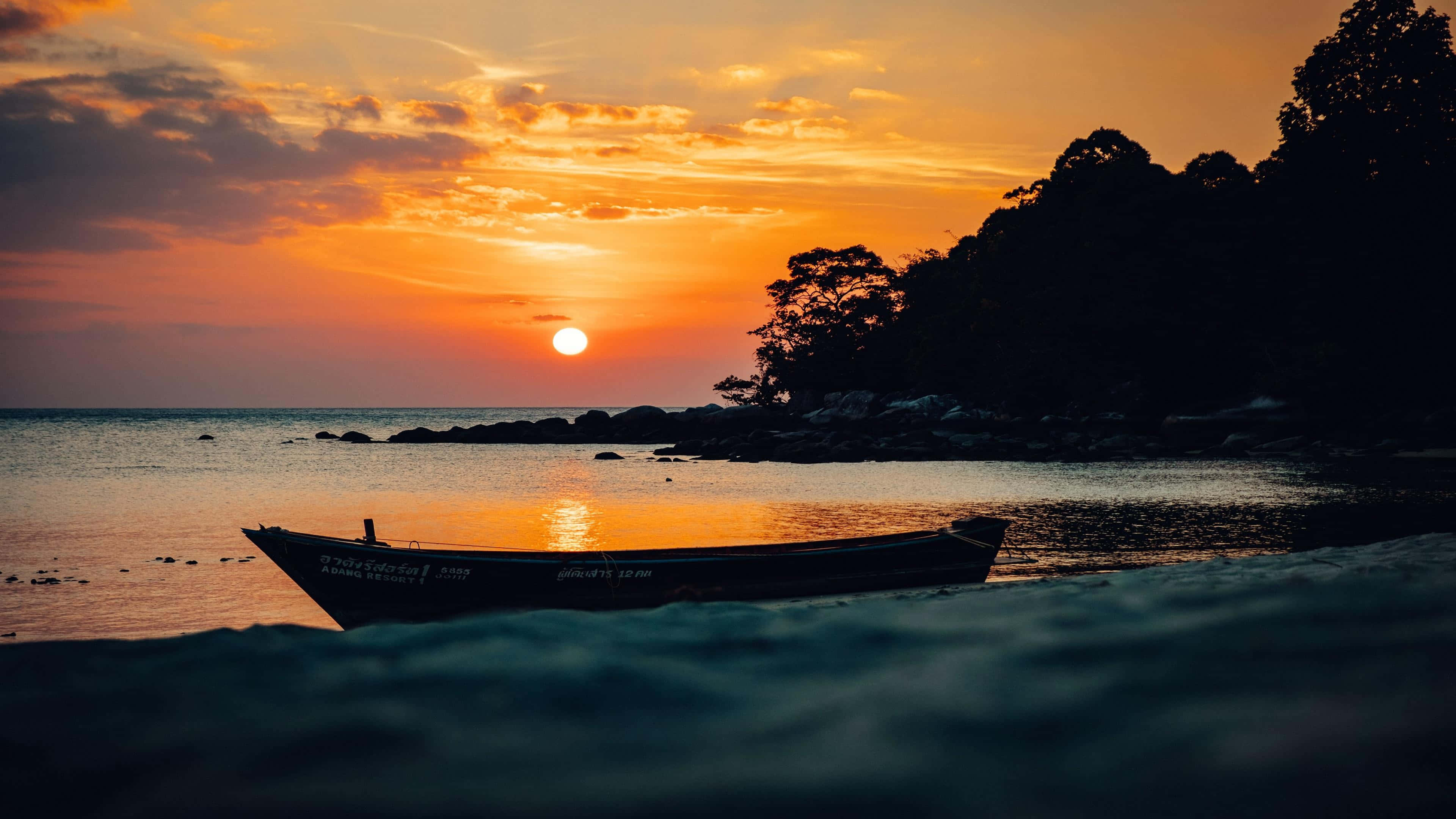 Seashore 桌布 壁紙 — 一艘孤獨的小船停泊在沙灘上，夕陽在樹影後落下，將溫暖的橙色光芒灑在平靜的水面上。