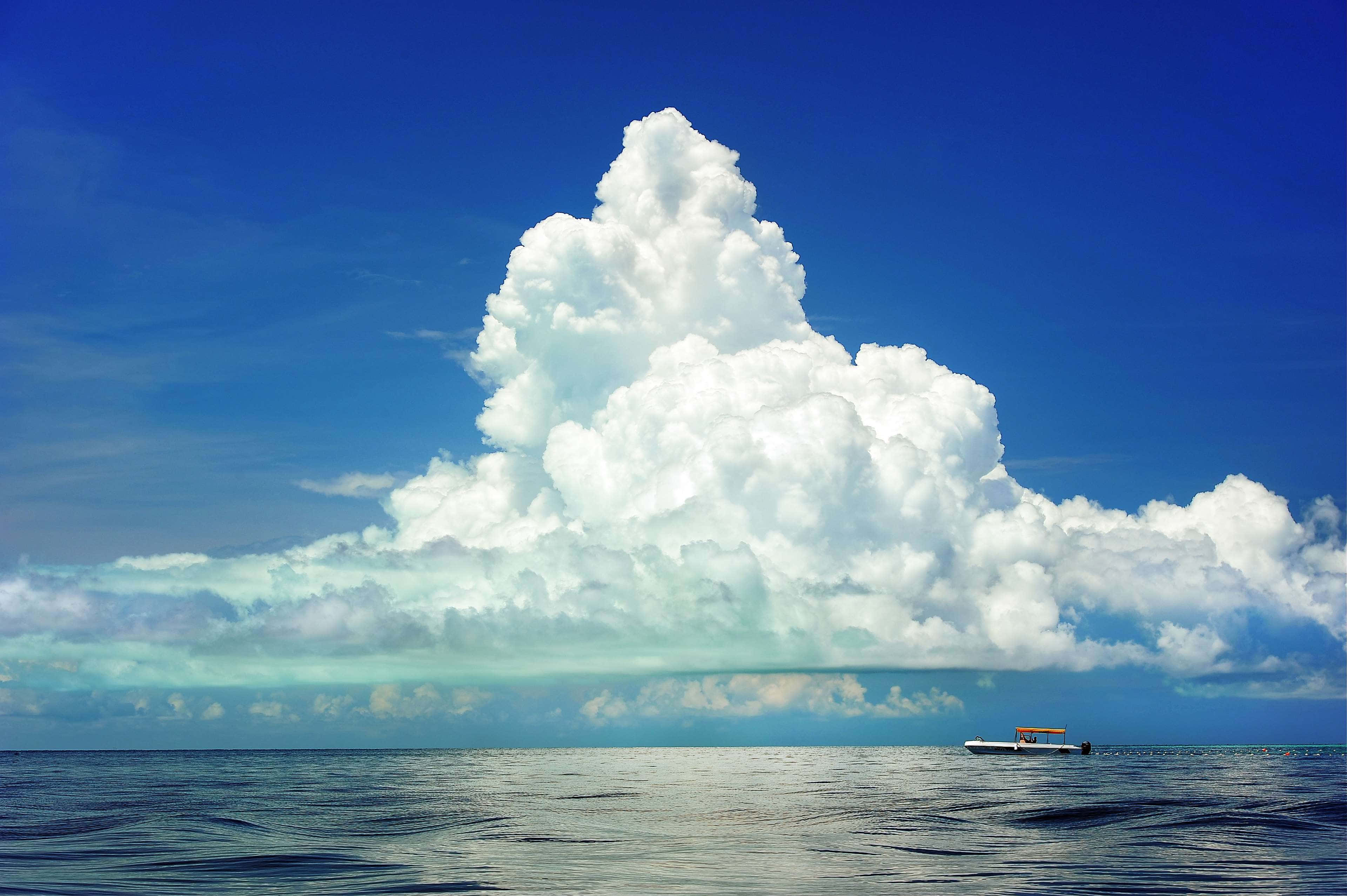 Boating 桌布 壁紙 — 一艘孤獨的小船漂浮在平靜的海面上，上方是巨大蓬鬆的白雲，映襯著鮮豔的藍天。