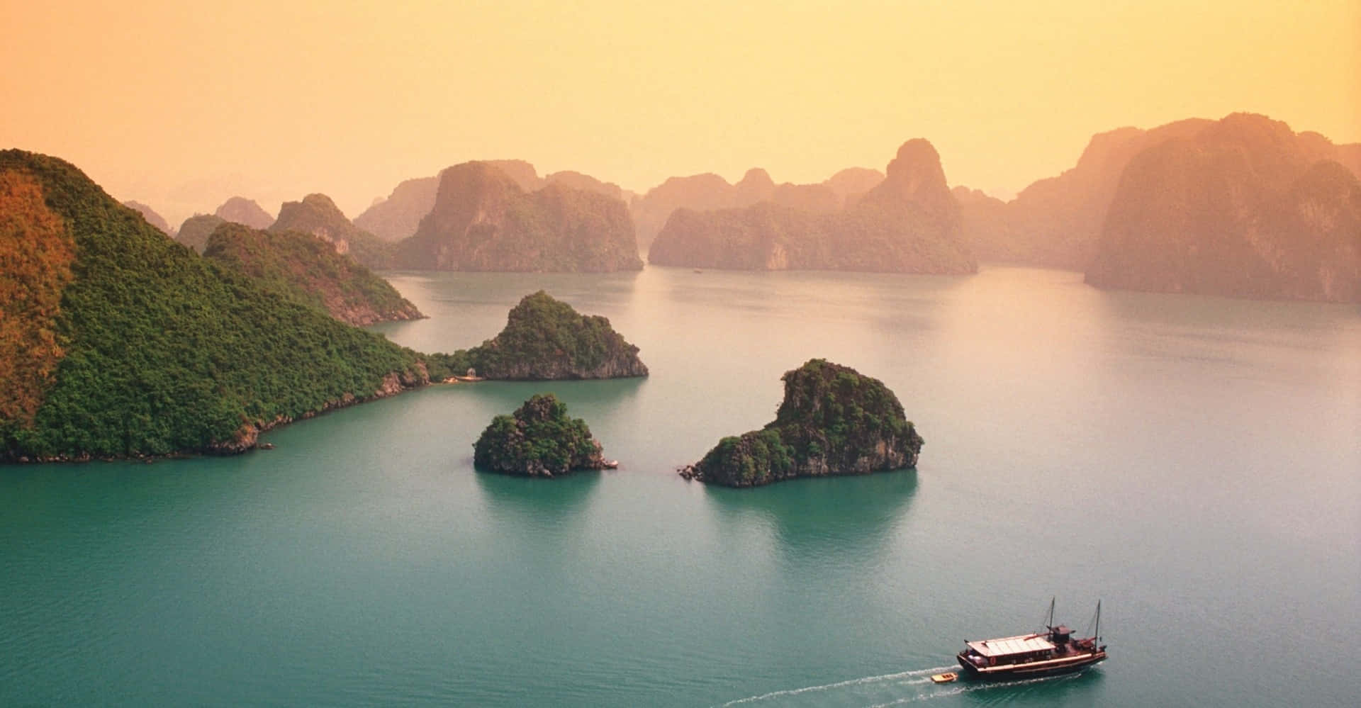 Vietnam 桌布 壁紙 — 一艘小船在溫暖的金色日落天空下，滑過碧綠海水，經過鬱鬱蔥蔥的綠色喀斯特島嶼。