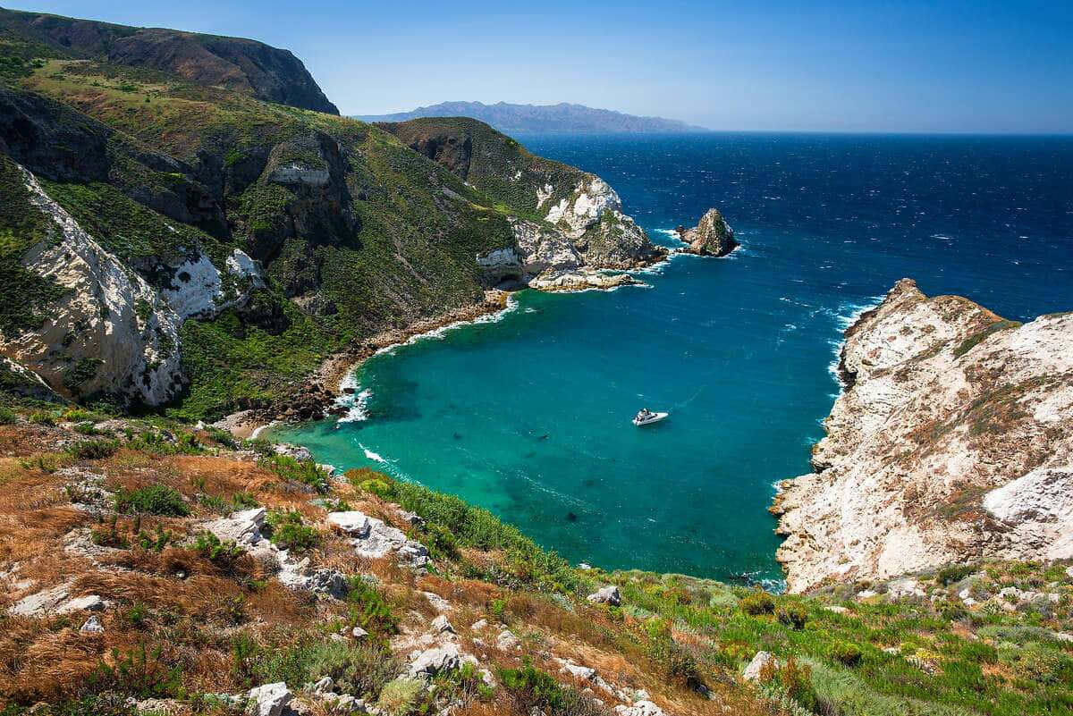 California Island 桌布 壁紙 — 一艘小船漂浮在碧綠色的海灣中，四周是崎嶇的綠色和白色懸崖，上方是清澈的藍天。