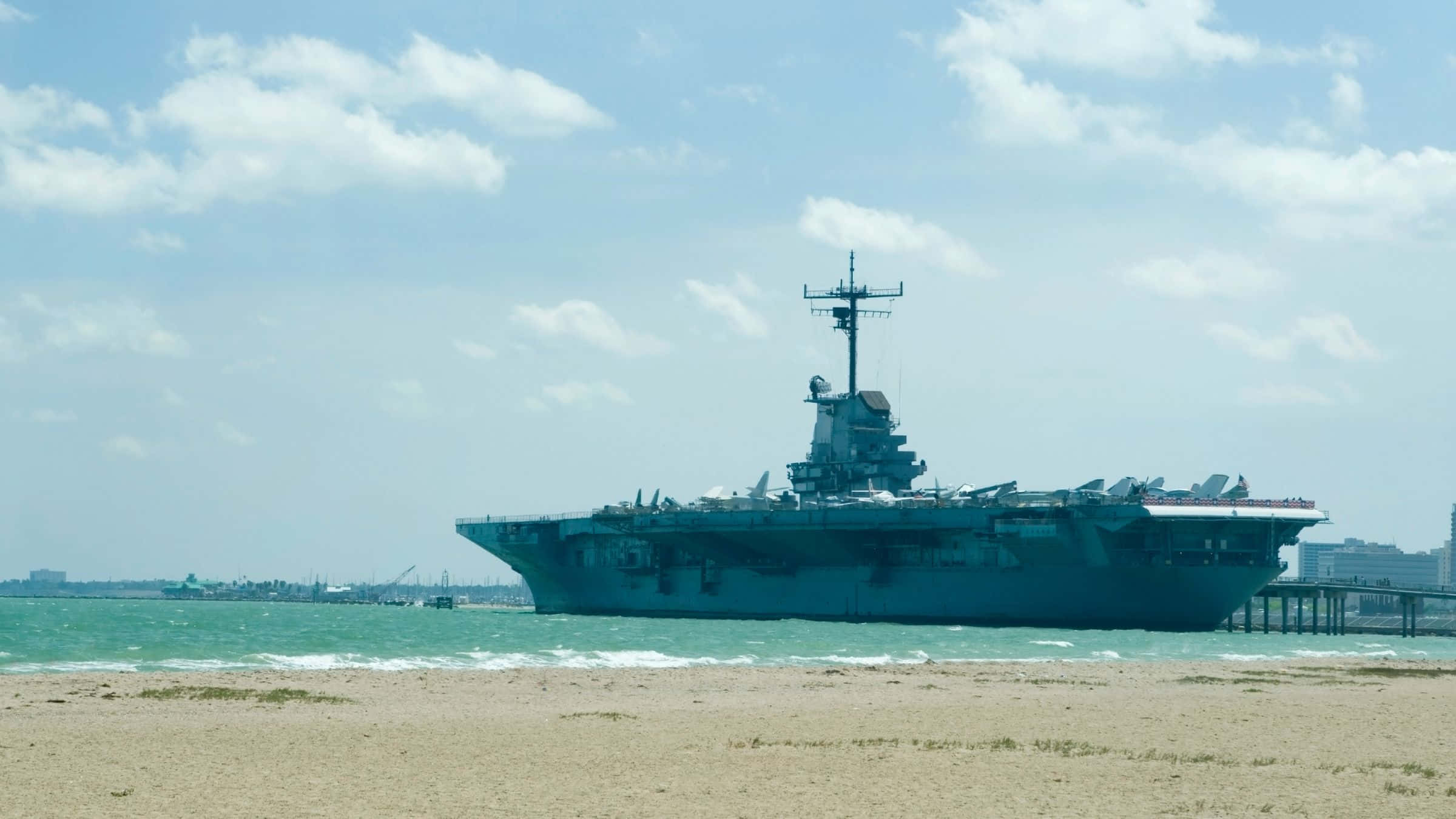 德州 桌布 壁紙 — 一艘巨大的灰色航空母艦停泊在沙灘附近平靜的碧綠海水上，天空部分多雲。