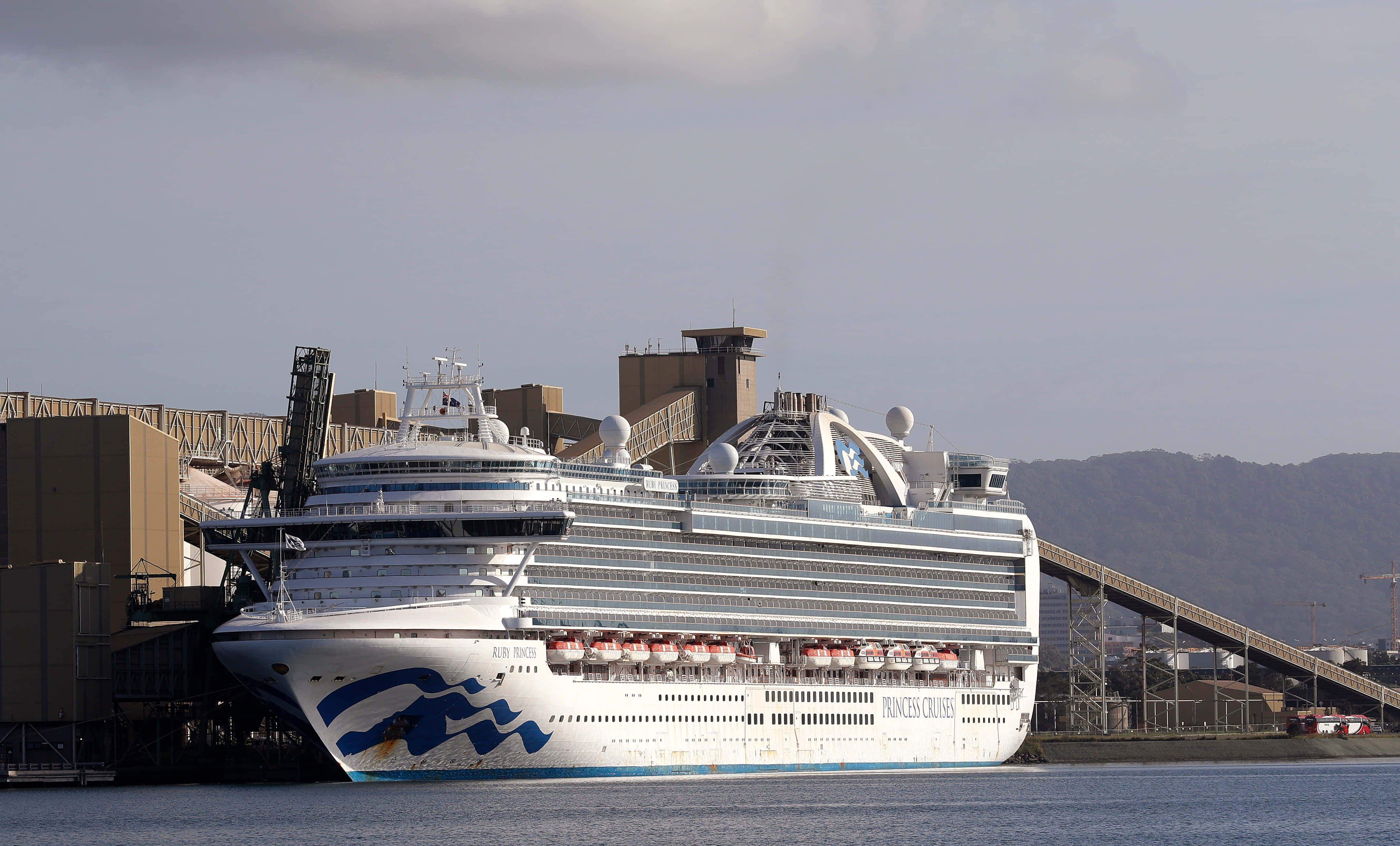 Cruise Ship 桌布 壁紙 — 一艘巨大的白色公主郵輪停泊在港口，背景是工業建築物和山丘，天空多雲。