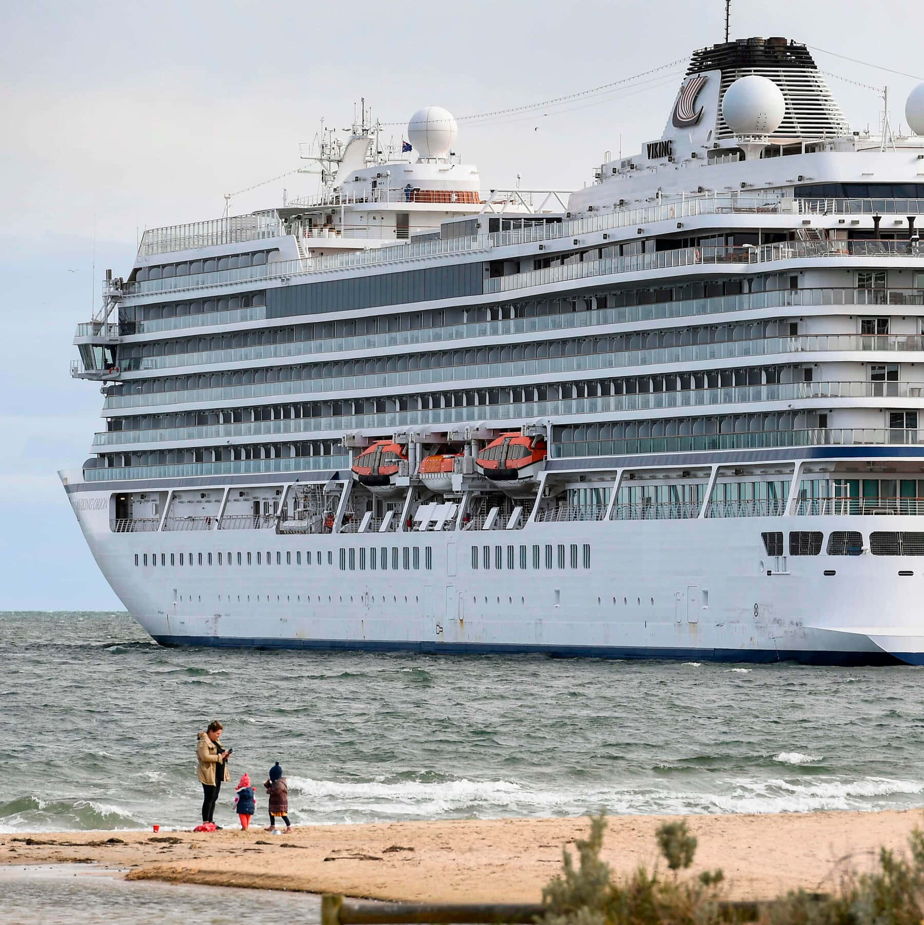 Cruise Ship 桌布 壁紙 — 一艘巨大的白色郵輪主宰著海洋，一名女子和兩個孩子站在沙灘上看著它經過。