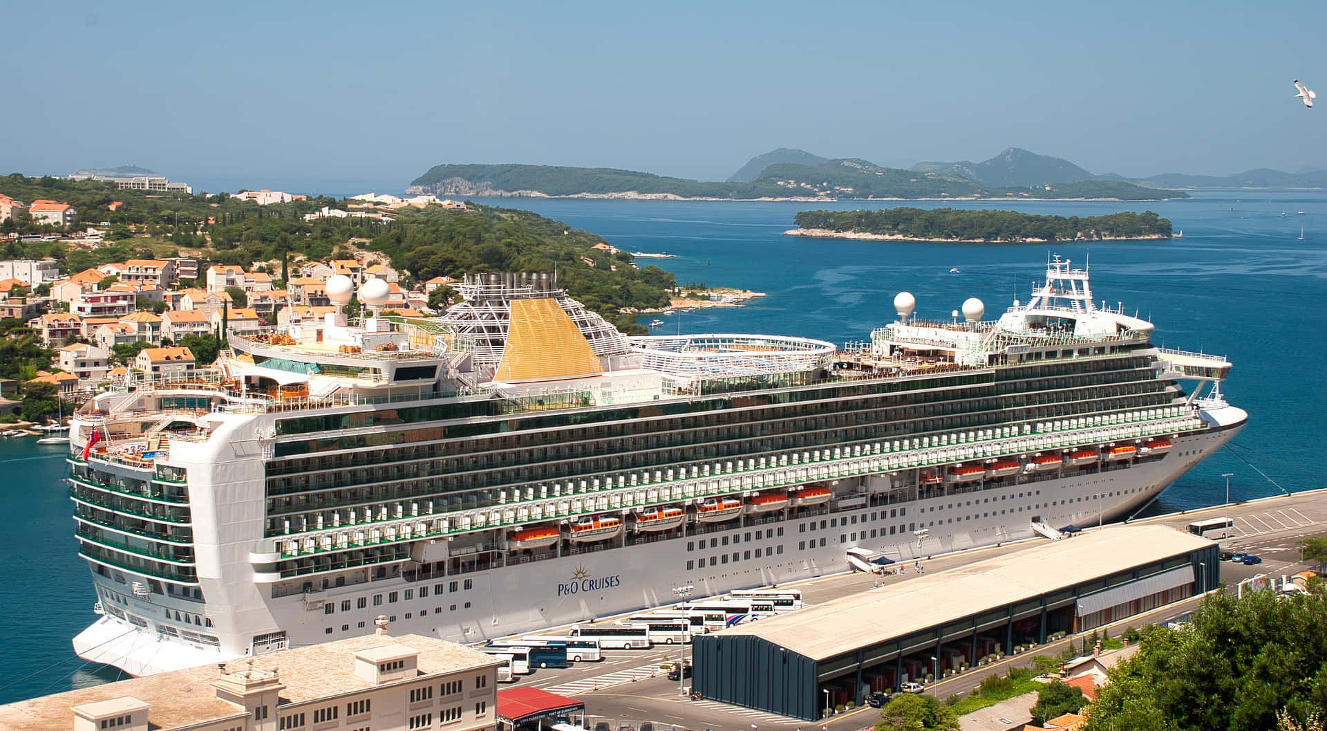 Cruise Ship 桌布 壁紙 — 一艘巨大的白色郵輪停泊在陽光明媚的港口，港口有陶土屋頂的建築物和延伸至綠色山丘的碧綠海水。